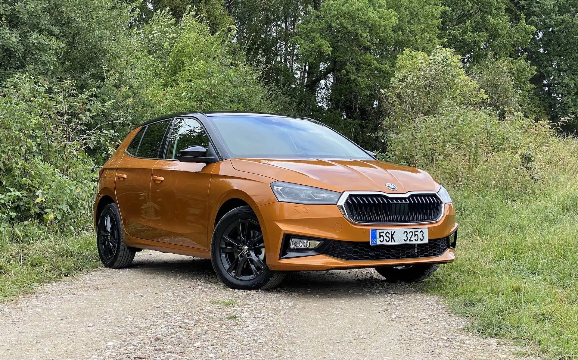 Perfil lateral completo de un moderno hatchback Skoda Fabia 2021 de color naranja estacionado en un camino sin pavimentar con vegetación en el fondo, capturado en un ángulo ligeramente oblicuo desde el frente, destacando el lado del conductor. Características incluyen una rejilla prominente con el emblema de la marca centrado, faros elegantes, luces antiniebla, grandes ruedas de aleación negras de varios radios, manijas de puertas y espejos color carrocería, línea de techo inclinada y un pequeño vistazo a la parte trasera y el conjunto de luces.