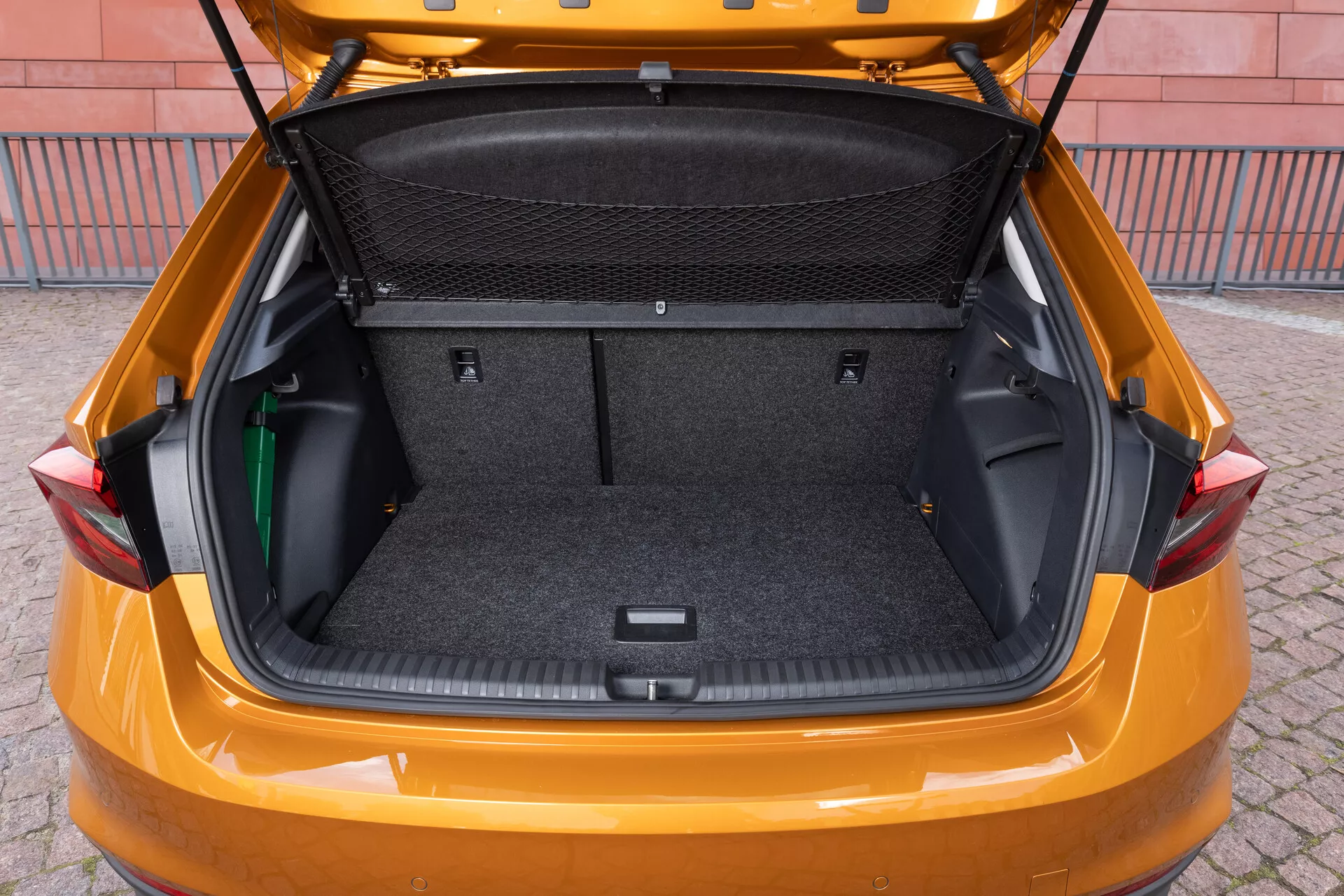 La imagen muestra el maletero de un modelo Skoda Fabia 2021, tomada desde un ángulo trasero ligeramente elevado para mostrar el espacioso área de carga. El interior está forrado con un material similar a una alfombra y cuenta con áreas de almacenamiento laterales flexibles, una partición de red para carga y una zona empotrada en la parte inferior para almacenamiento adicional. La compuerta está levantada, revelando un maletero grande y limpio, diseñado para resaltar la capacidad de almacenamiento y la practicidad del vehículo.