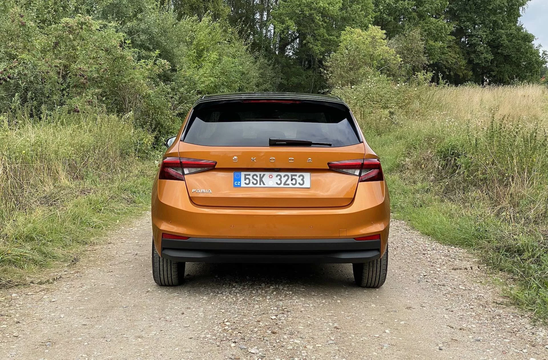 Vista trasera de un Skoda Fabia 2021 hatchback de color naranja metálico brillante fotografiado a la luz del día en un camino de tierra estrecho en un entorno rural. La imagen captura el diseño trasero moderno con una barra de luz continua, luces traseras angulares, reflectores integrados, limpia parabrisas trasero y spoiler trasero para aerodinámica, destacando el acabado brillante y las características prácticas del coche.