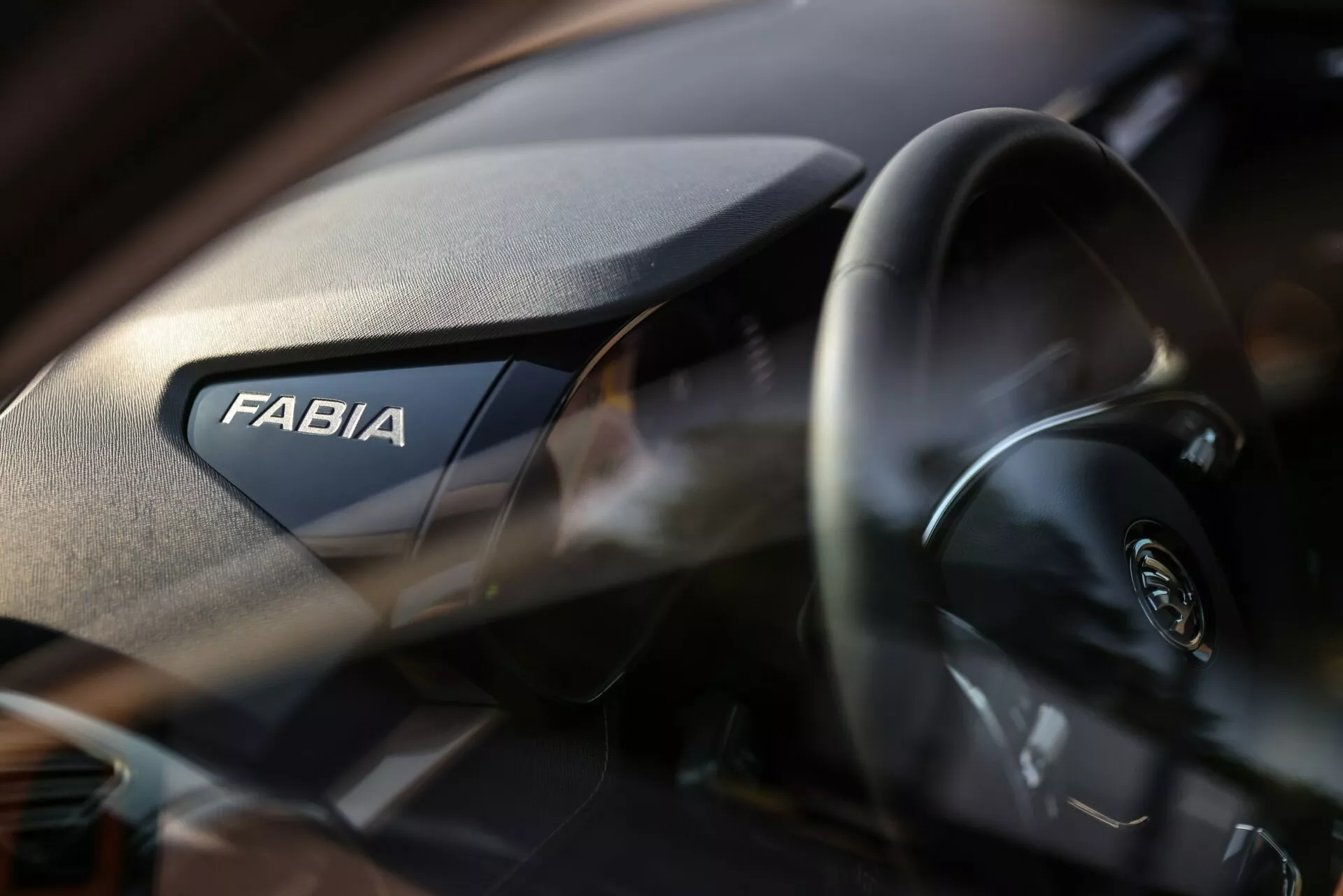Vista interior de cerca del Skoda Fabia 2021, destacando el lado del pasajero del tablero. La palabra 'FABIA' se muestra de manera prominente, mientras que el volante moderno con el logotipo de Skoda es parcialmente visible a la izquierda. El tablero tiene una textura granulada y la poca profundidad de campo enfatiza la marca con un fondo suavemente desenfocado.