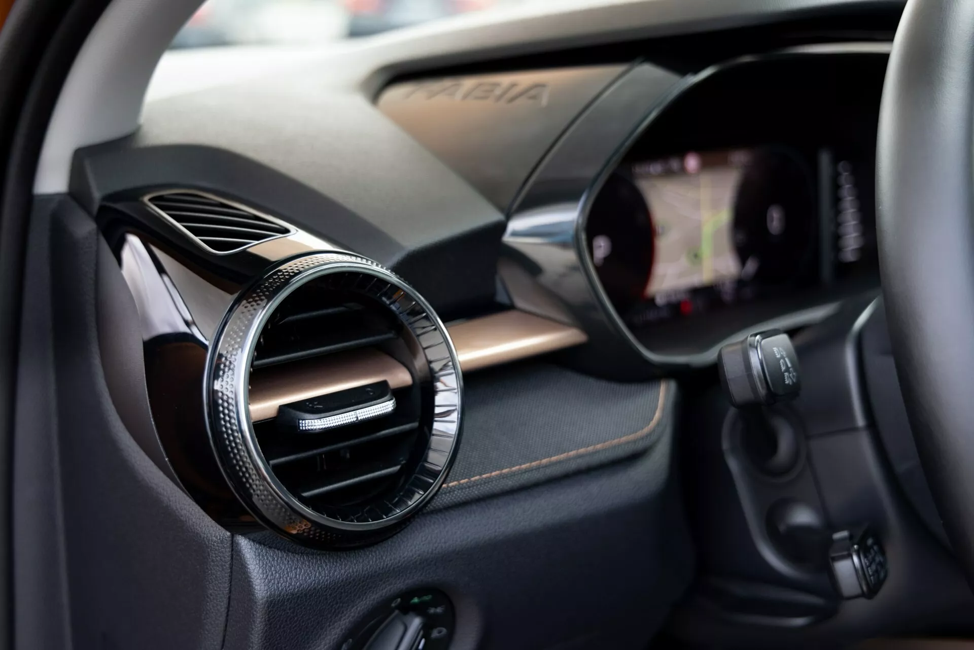 Vista en primer plano del interior del Škoda Fabia 2021 centrada en el lado del pasajero. La imagen captura una salida de aire redonda con un acabado metálico o cromado contra un panel texturizado como fibra de carbono, indicando un acabado deportivo o de alta gama. A la derecha está visible el borde de una pantalla táctil de infoentretenimiento integrada en el tablero. Debajo de la salida de aire hay varios botones de control, posiblemente para el sistema de audio, el clima u otras funciones del vehículo. La poca profundidad de campo llama la atención sobre la salida de aire, mientras sugiere suavemente los componentes tecnológicos circundantes.