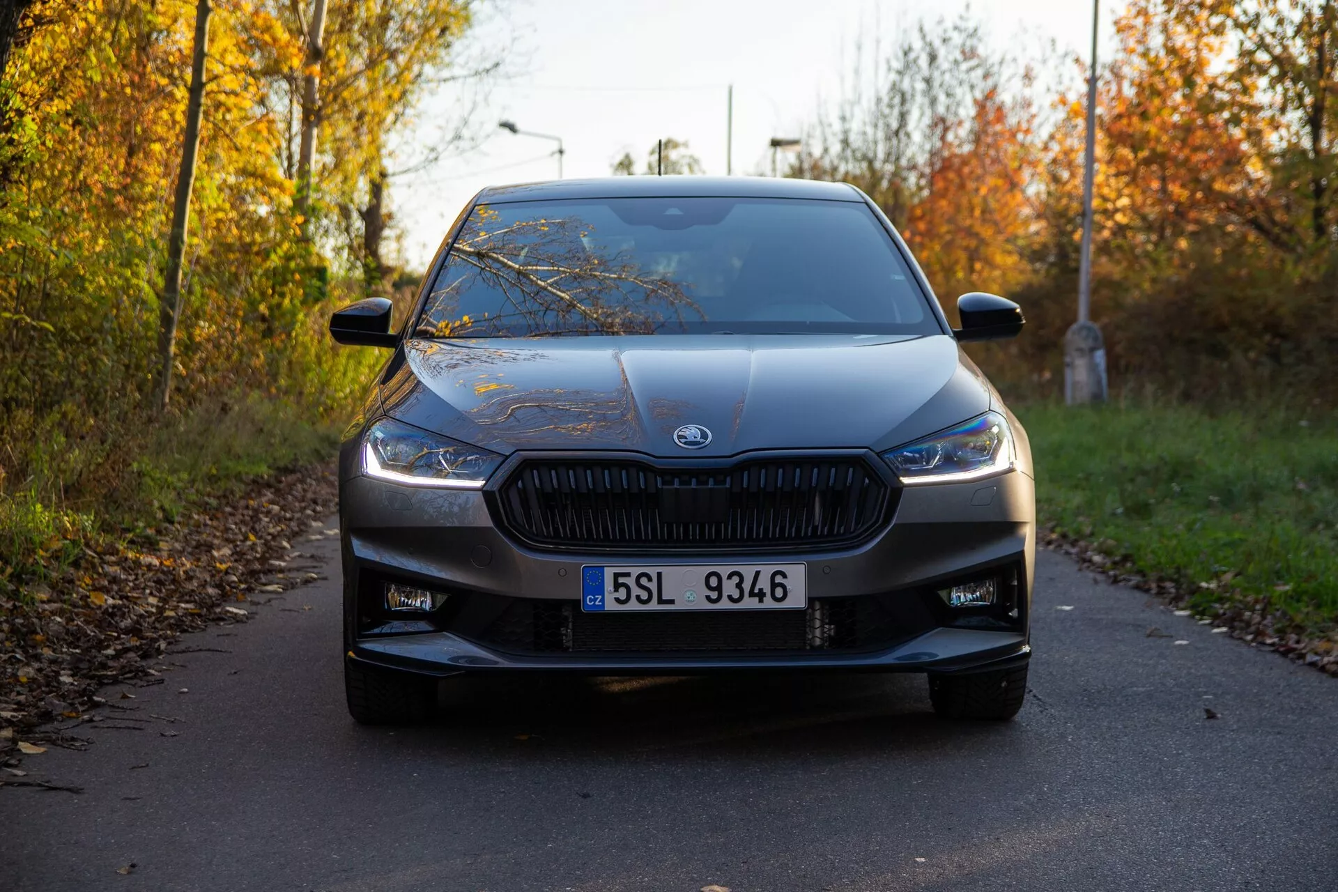 Vista frontal de un Skoda Fabia 2021 capturada desde un ángulo bajo, destacando su distintiva parrilla con listones verticales y faros modernos y estrechos. El coche, centrado en el marco, está estacionado en una carretera estrecha rodeada de follaje otoñal, adornado con el emblema del fabricante prominentemente visible en el centro de la parrilla. Otros detalles de diseño visibles incluyen la matrícula clara, tomas de aire en el parachoques inferior, líneas de contorno suaves en el capó, parabrisas expansivo y espejos laterales con indicadores. La suave iluminación y las largas sombras sugieren que la imagen fue tomada por la mañana temprano o al caer la tarde, mostrando el coche en excelentes condiciones.