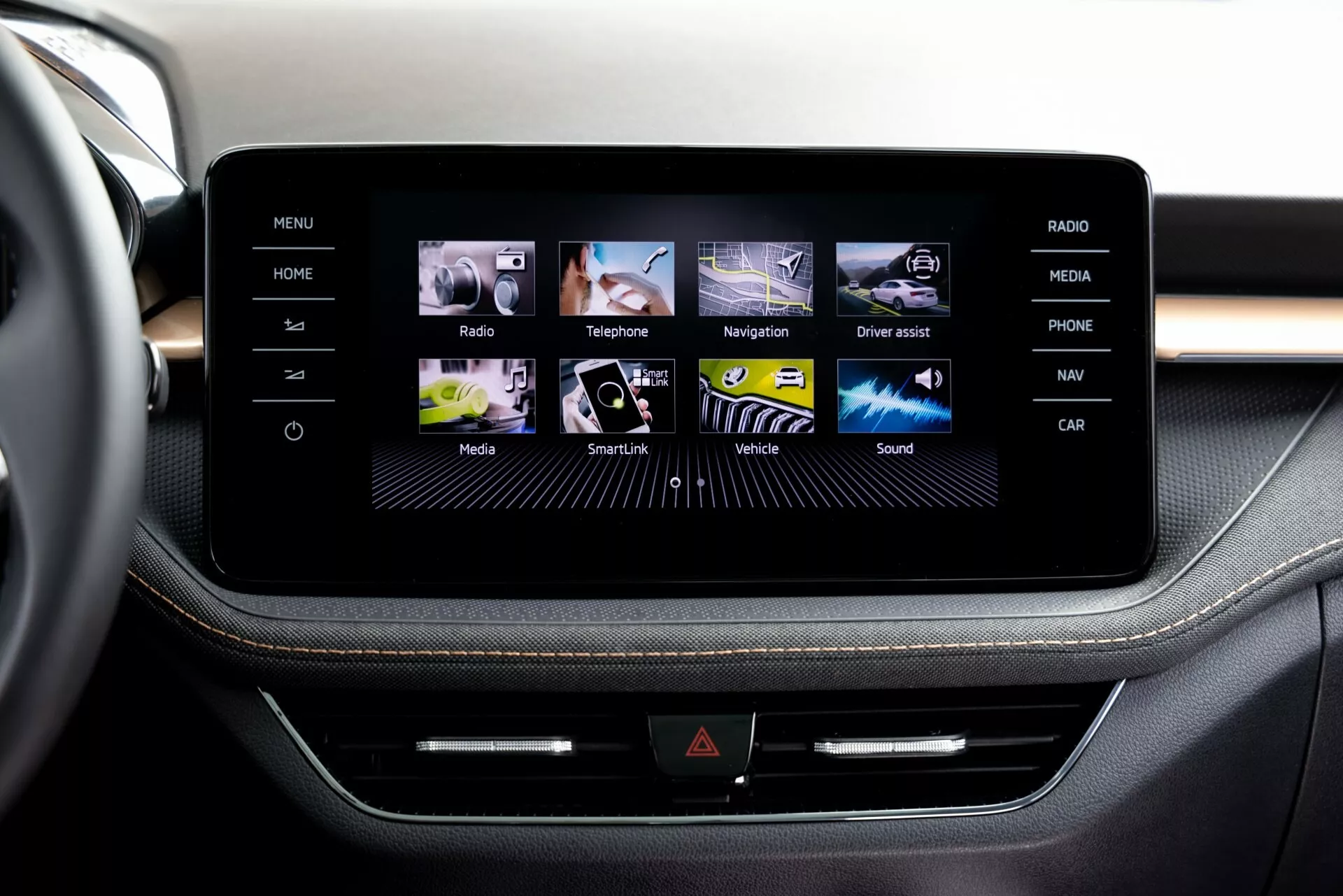 Vista de cerca del sistema de infoentretenimiento con pantalla táctil del Škoda Fabia 2021 desde la perspectiva de un pasajero o conductor en la fila delantera, mostrando una consola central elegante enmarcada en lo que parece ser aluminio cepillado. La pantalla muestra una interfaz gráfica amigable con íconos coloridos para Home, Radio, Teléfono, Navegación y más. Debajo de la pantalla, una línea de botones físicos y una posible ranura para CD añaden multifuncionalidad. La tapicería premium con costuras contrastantes sugiere un diseño interior de alta gama.