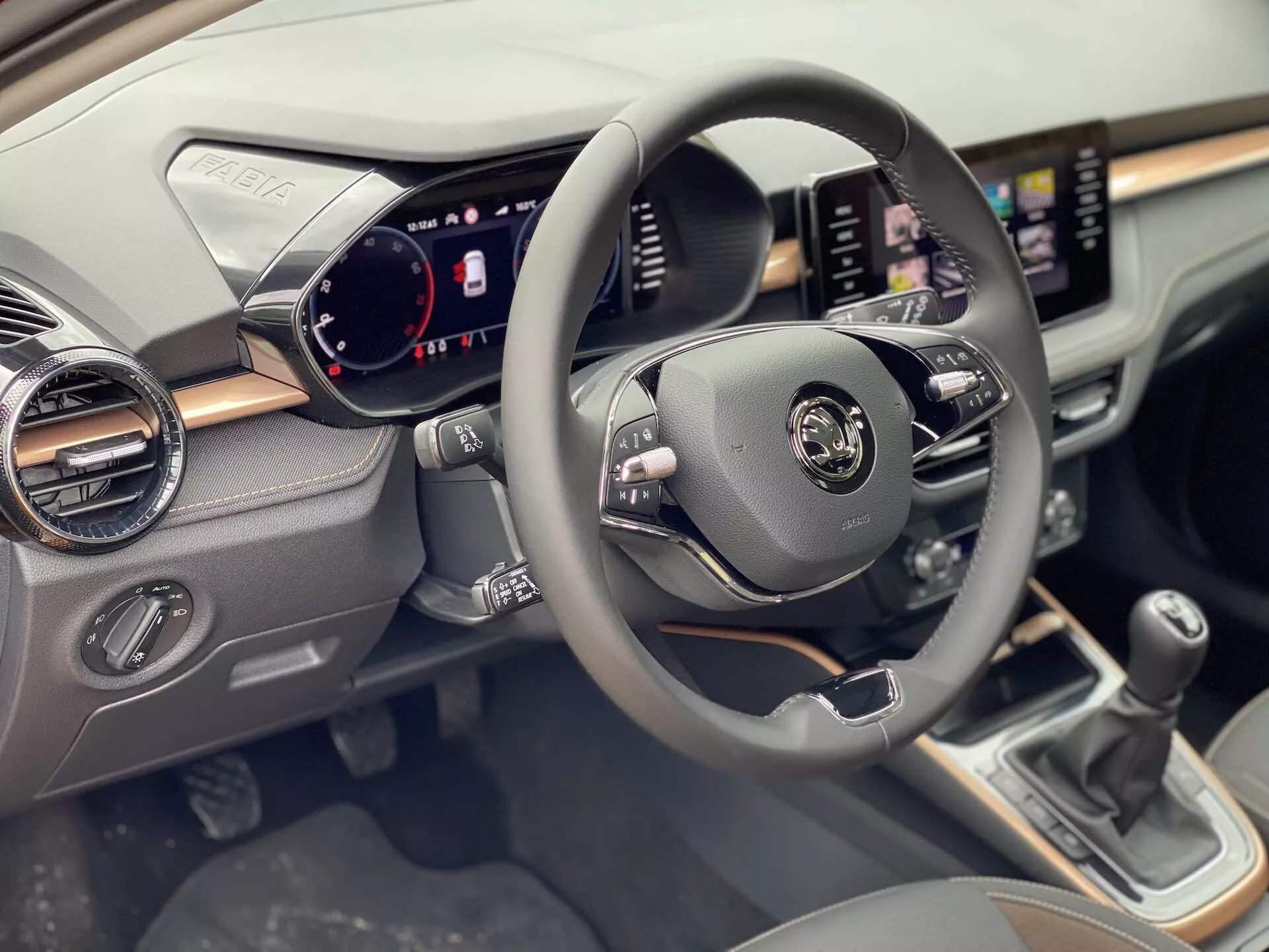 Vista interior del Skoda Fabia 2021 enfocada en el lado del conductor, mostrando el volante centrado con controles multifuncionales para audio, comandos de voz y control de crucero. El panel de instrumentos digital muestra velocímetro, tacómetro e indicadores de estado del vehículo. Un sistema de infoentretenimiento con pantalla táctil se muestra en el tablero con funcionalidades de navegación y medios. Debajo de la pantalla hay diales y botones de control climático. La palanca de cambios manual es visible en la consola central.