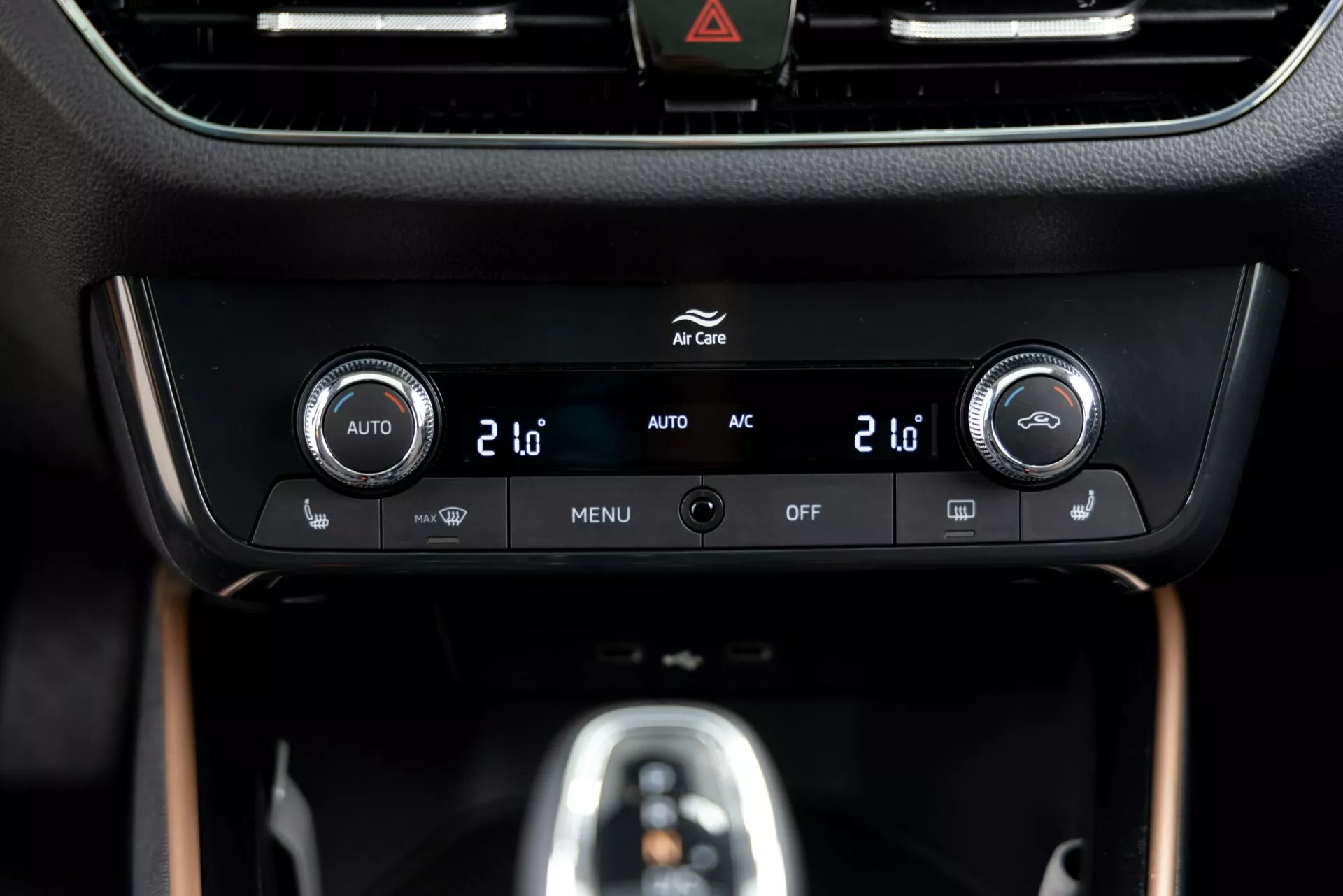 Vista en primer plano del panel de control climático del Skoda Fabia 2021 ubicado en la consola central, con controles de temperatura de doble zona con pantallas digitales que muestran '20', botones integrados para el control climático automático ('AUTO'), aire acondicionado ('AC'), configuraciones del menú ('MENU') y apagado del sistema ('OFF'). Un borde negro brillante rodea una pantalla de información encima de los controles, con un botón de advertencia de peligro (icono de triángulo) visible en la parte superior. El panel está integrado en un interior premium con acabado mate y de apariencia de madera, destacando un diseño y tecnología modernos.