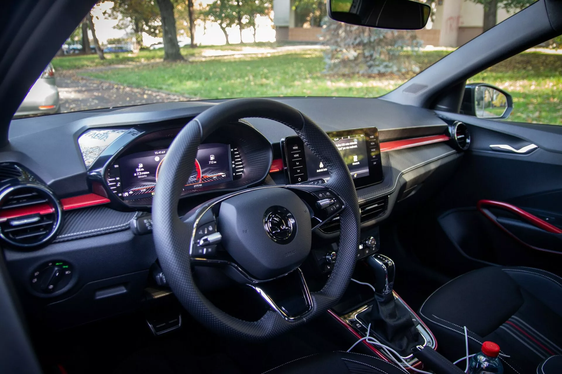 Vista interior de un moderno Skoda Fabia 2021 desde la perspectiva del conductor, con un panel de control en la puerta del conductor, un volante multifuncional, un cuadro de instrumentos digital, salidas de aire con acentos rojos, una pantalla táctil central de infoentretenimiento, una palanca de cambios manual y el área del asiento del pasajero con una guantera y una salida de aire a juego.