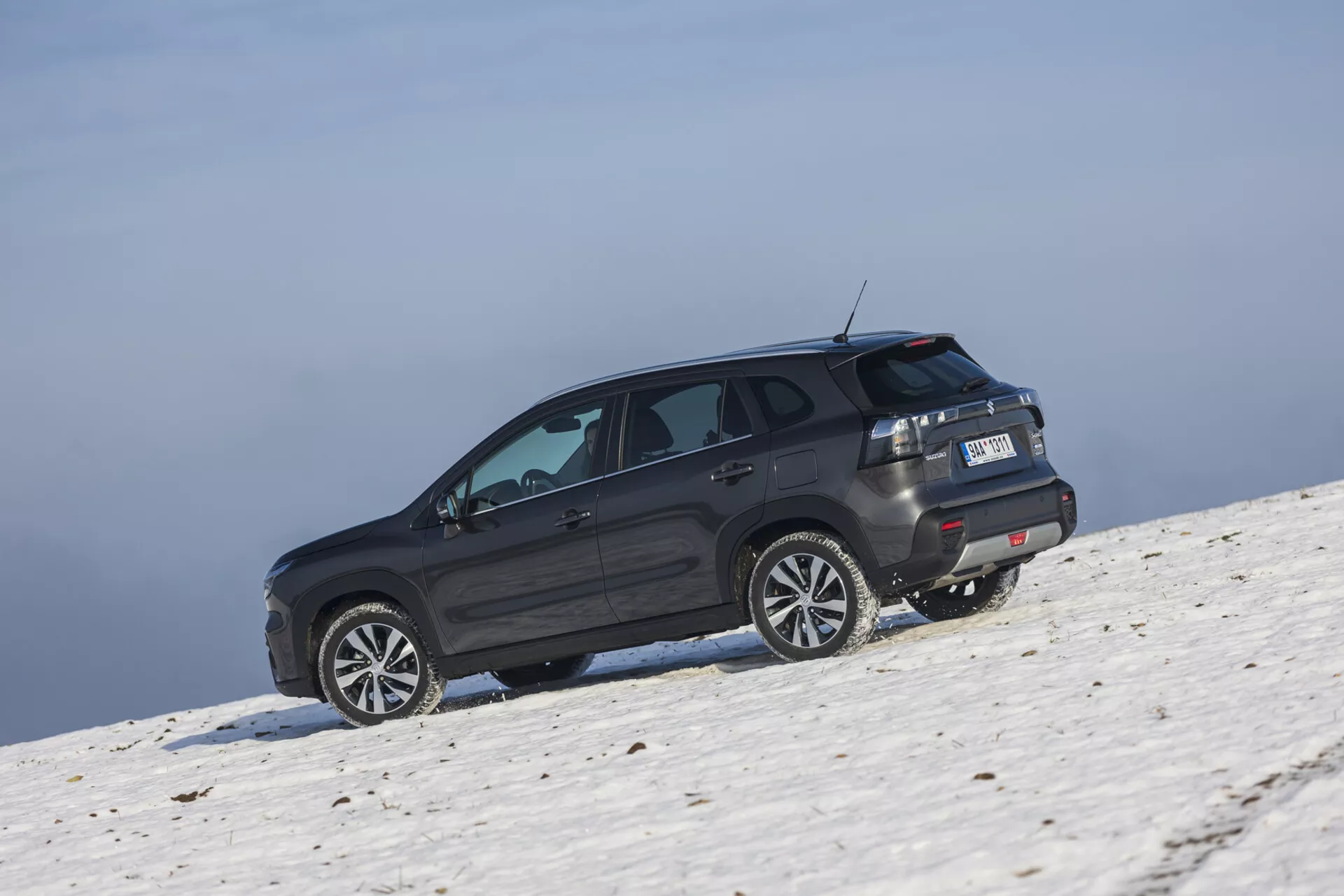 SUV Suzuki S-Cross 2022 en un tono oscuro posicionado en una pendiente nevada capturado desde un ángulo trasero oblicuo resaltando su perfil lateral y trasero, llantas de aleación grandes, ventanas tintadas con desempañador, luces traseras LED, spoiler trasero, y capacidades potenciales fuera de carretera bajo condiciones de cielo claro.
