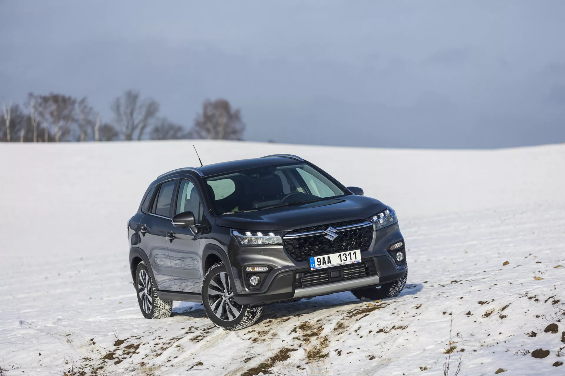SUV compacto Suzuki S-Cross de color oscuro del 2022 estacionado en un paisaje nevado, mostrando una vista frontal y lateral a un ángulo de 45 grados, exhibiendo una parrilla elegante con acabado metálico, faros LED alargados, ruedas de aleación de múltiples radios, y un borde de ventana cromado pulido indicando un paquete de lujo y potencial capacidad fuera de carretera