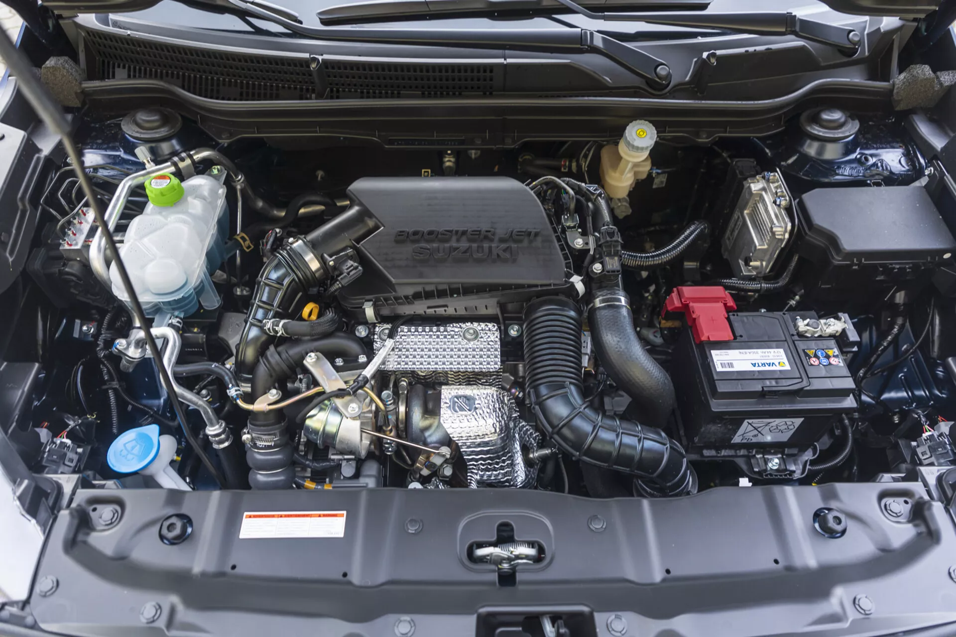 Bahía del motor del Suzuki S-Cross 2022 mostrando una vista frontal con componentes visibles como el bloque del motor, la cabeza del cilindro, el colector de admisión y características tecnológicas incluyendo el turbocompresor y el control de tiempo de válvulas variable, junto con la marca Suzuki en la cubierta del motor, en perfectas condiciones.