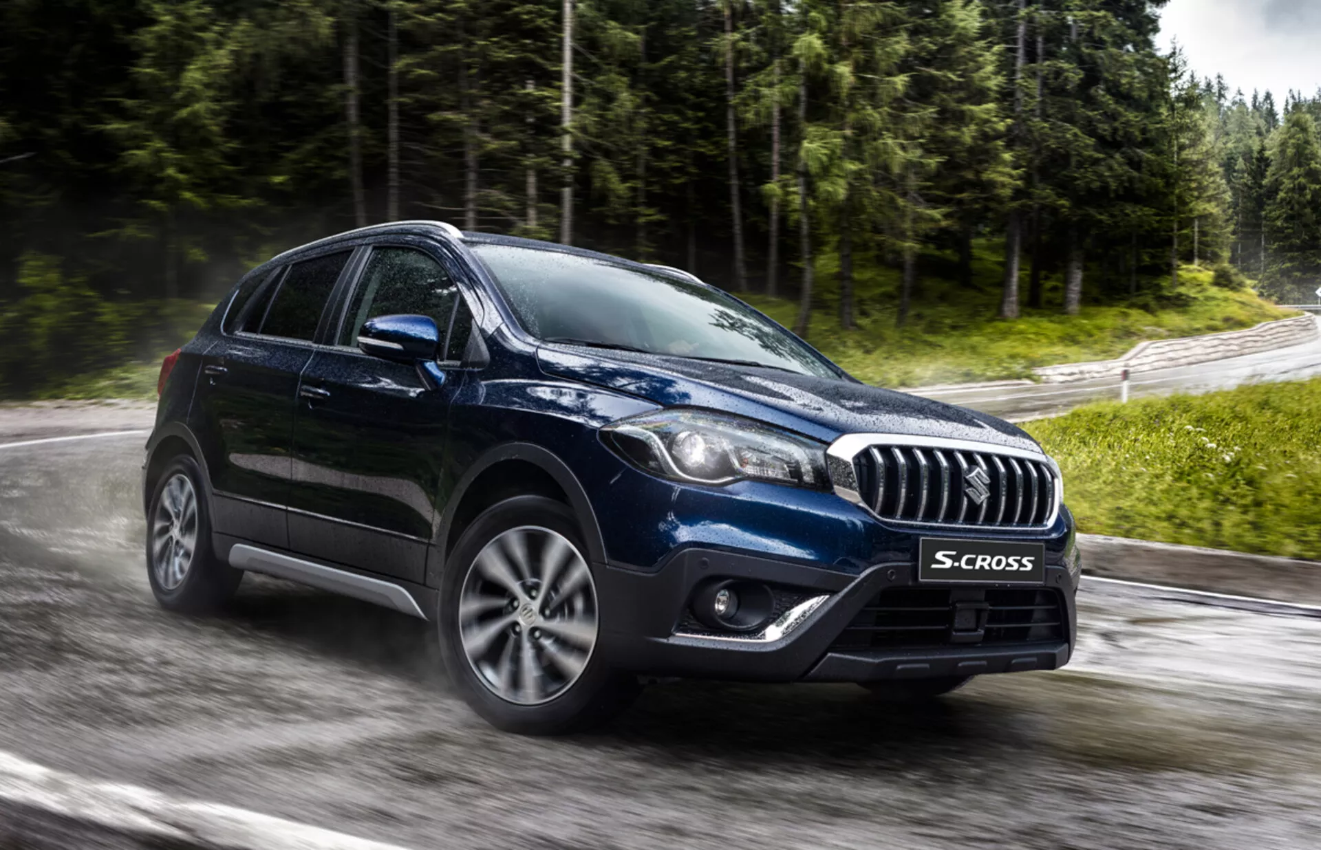 Suzuki S-Cross 2022 capturado desde un ángulo frontal de tres cuartos destacando su parrilla acentuada en cromo, faros modernos LED o HID, ruedas de aleación de múltiples radios, y forma robusta de crossover en una carretera mojada y rodeada de vegetación, sugiriendo tecnología avanzada y capacidad para todo tipo de clima.