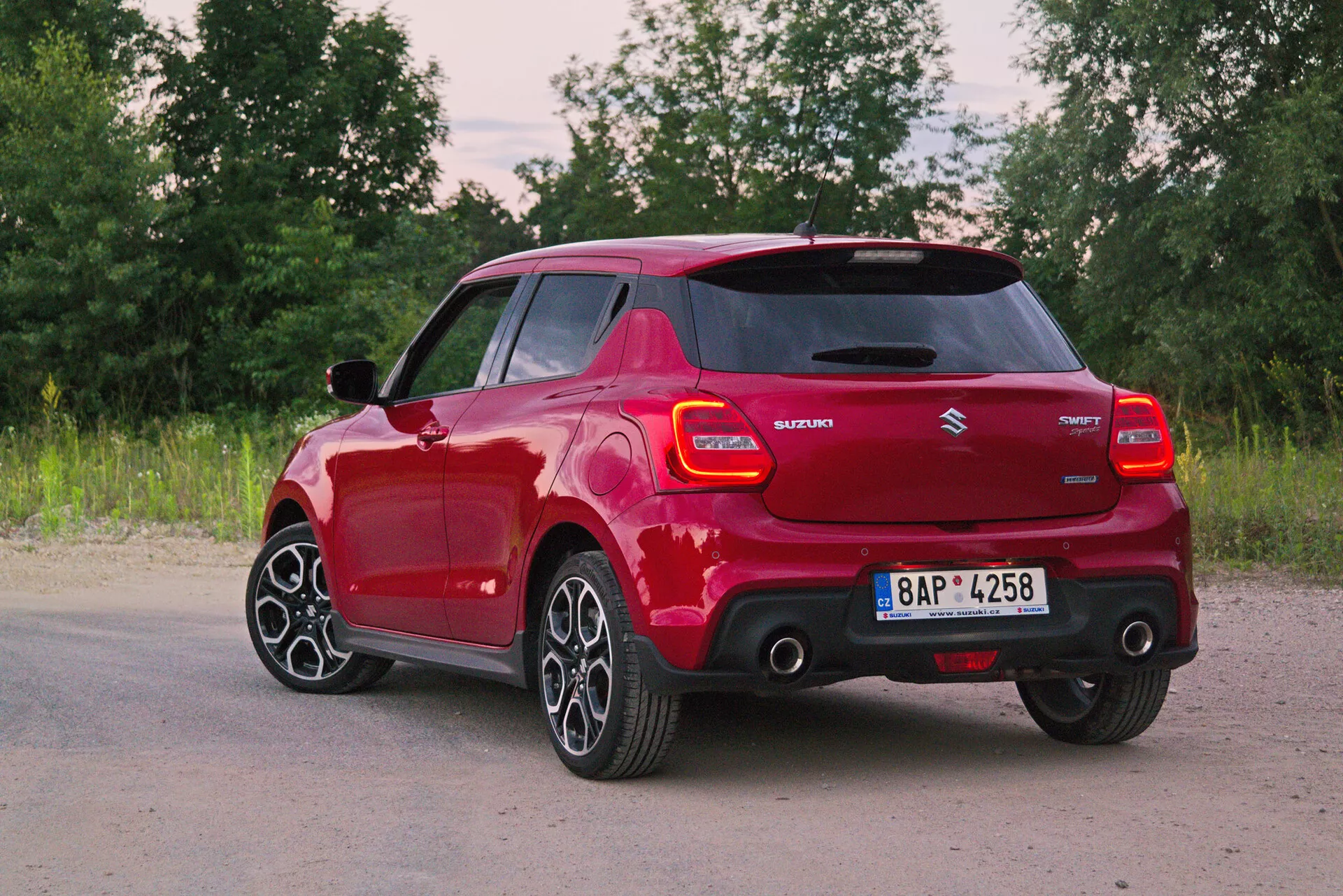 Imagen de un Suzuki Swift rojo 2017 Swift-Sport en vista tres cuartos trasera, mostrando elementos de diseño modernos como luces traseras LED, spoiler integrado, antena de aleta de tiburón, llantas de aleación multirradio, manijas de puertas y espejos laterales del color de la carrocería, y distintivos de tecnología híbrida. El fondo natural añade una sensación serena y dinámica.