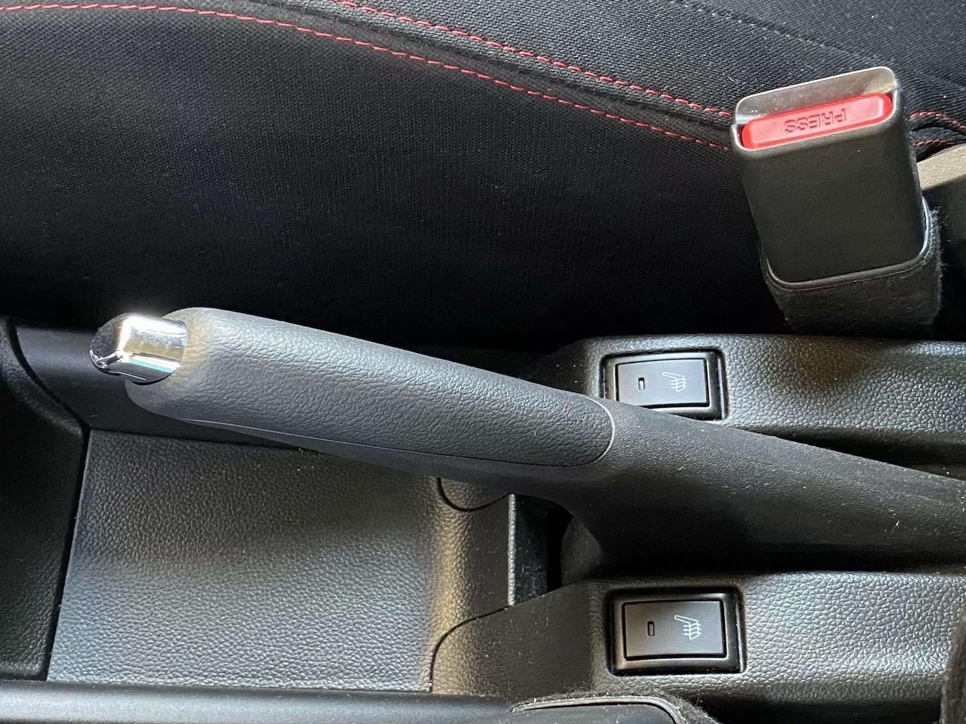Vista de primer plano del interior del Suzuki Swift 2017 (Swift Sport) que se enfoca en la palanca del freno de mano de cuero con un botón metálico y los controles de calefacción de los asientos. La palanca del freno de mano está centrada y ligeramente inclinada a la izquierda, indicando que está activada. Dos interruptores de botón con indicadores LED y un icono de asiento, ubicados al lado del freno de mano, sugieren calefacción de doble zona para los asientos delanteros. Un broche del cinturón de seguridad con botón de liberación rojo es parcialmente visible detrás de los controles. La imagen está tomada desde un ángulo elevado con iluminación diurna natural, destacando las características de tecnología y confort.