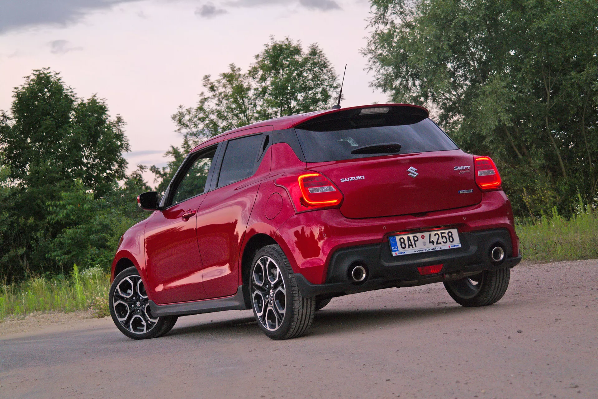 Hatchback rojo Suzuki Swift Sport 2017 capturado desde un ángulo de tres cuartos trasero, destacando su perfil trasero y lateral del lado del conductor. Foto tomada desde una perspectiva ligeramente baja, enfatizando las modernas ruedas de aleación de múltiples radios y neumáticos de perfil bajo. Luz natural suave proporcionada por condiciones de amanecer o atardecer con nubes parciales. Las características visibles incluyen el diseño contemporáneo de las luces traseras con elementos LED, la insignia del fabricante, el nombre del modelo, espejos laterales del color de la carrocería posiblemente con indicadores de giro, parachoques trasero con sensores de estacionamiento integrados y una línea del techo que sugiere un alerón para eficiencia aerodinámica.