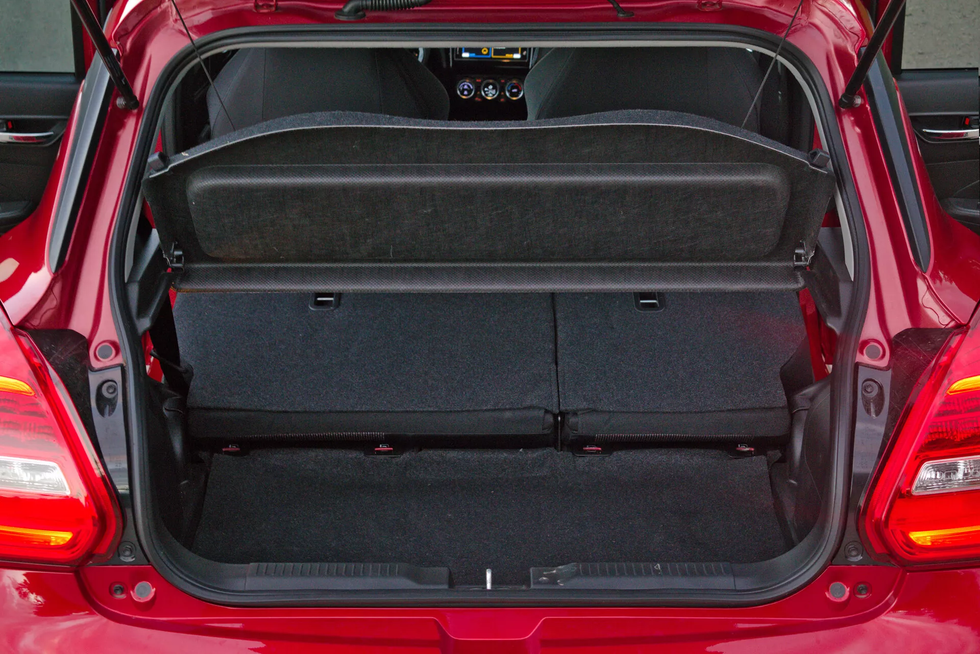 Espacio de maletero trasero de un Suzuki Swift Sport 2017 hatchback con la puerta trasera abierta, mostrando área de carga vacía con alfombra automotriz oscura. Vista desde la perspectiva trasera directa, revelando estante desmontable y plegable, y asientos traseros 60/40. Vista parcial del limpiaparabrisas trasero y componentes interiores, incluidos altavoz trasero y sistema de iluminación.