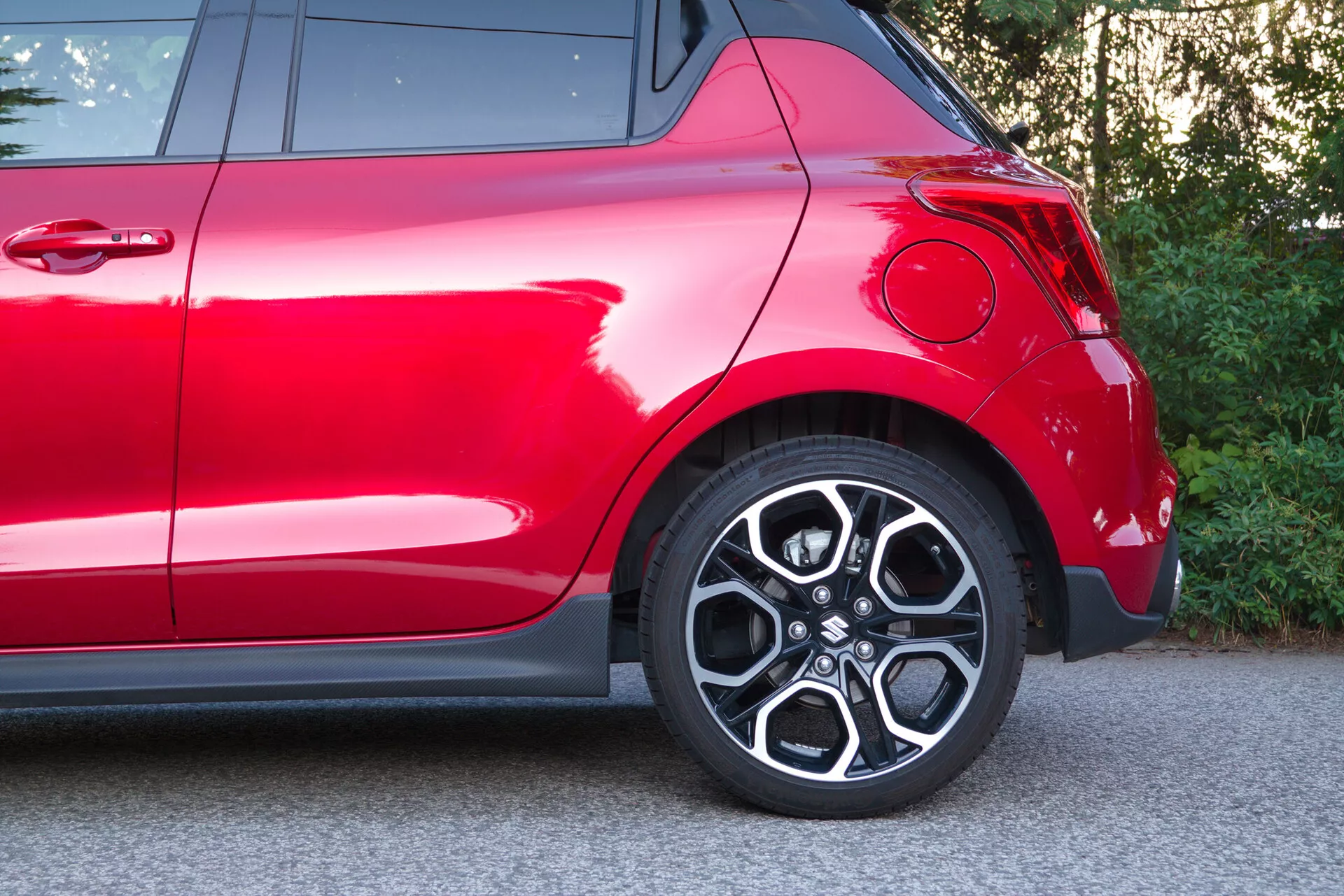 Vista lateral parcial de un Suzuki Swift 2017 (Swift Sport) rojo, enfocándose en la parte trasera, incluyendo la rueda y el área del guardabarros trasero. El enfoque captura la rueda de aleación elegante con diseño de múltiples radios, neumático de perfil bajo, guardabarros trasero con curva moderna, pintura roja profunda y brillante, parte de la luz trasera y elementos decorativos cromados, destacando su diseño deportivo y acabado exterior premium.