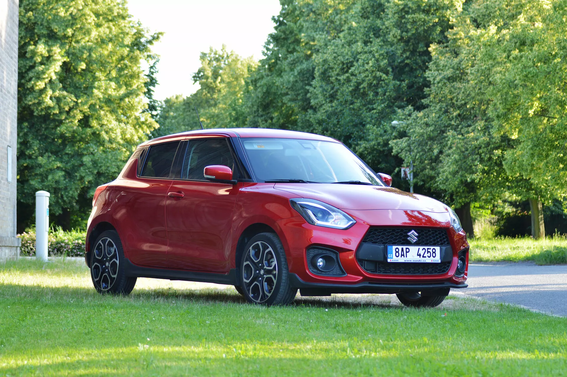 La imagen muestra un hatchback Suzuki Swift Sport rojo de 2017 ubicado en un área de césped, probablemente en un parque o al borde de la carretera, capturado desde un ángulo de tres cuartos frontal. La prominente parrilla delantera, los elegantes y angulares faros, la asamblea de luces antiniebla y el capó inclinado hacia un parabrisas aerodinámico se destacan. Las llantas de aleación, los espejos laterales del color de la carrocería, la línea del techo que desciende suavemente hacia la ventana trasera y las ventanas polarizadas contribuyen al diseño del vehículo. La iluminación natural y la vista despejada enfatizan la postura y el diseño general del coche.