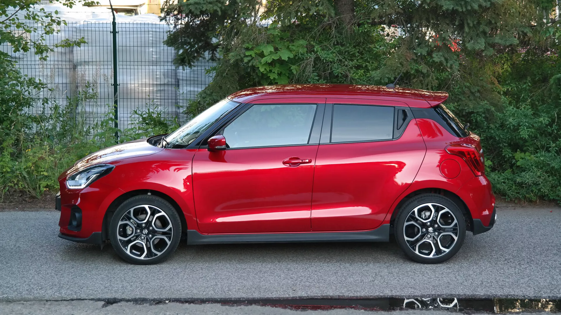Vista de perfil lateral de un coche hatchback Suzuki Swift Sport 2017 rojo. El vehículo se capta con iluminación natural diurna, mostrando las características de su elegante diseño, incluyendo la integración de los faros en el arco de la rueda delantera, llantas de aleación con múltiples radios, espejos laterales y manijas del color de la carrocería, un sistema de entrada sin llave, y un techo solar visible. El coche también cuenta con un alerón en el techo y luces traseras montadas en lo alto. La imagen, ambientada en una calle suburbana, enfatiza la aerodinámica del coche, sus características deportivas y probablemente sus avanzadas tecnologías de seguridad.