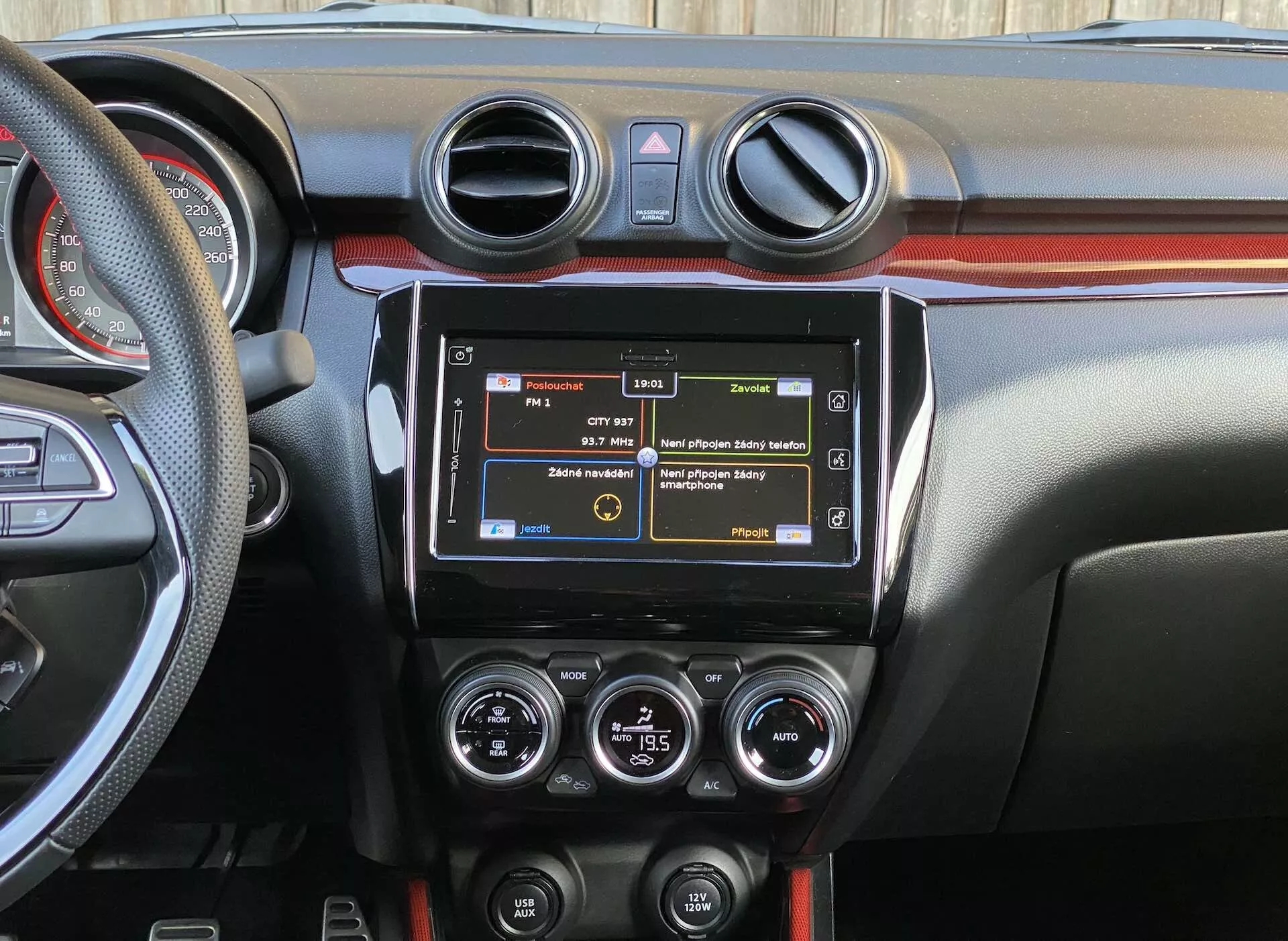Vista detallada del interior del Suzuki Swift Sport 2017 enfocada en el tablero central desde la perspectiva del asiento trasero, mostrando el sistema de infoentretenimiento moderno, ventilaciones circulares del aire acondicionado, controles de HVAC, varios botones de funciones, diseño de tablero bicolor y una parte del volante con controles montados.