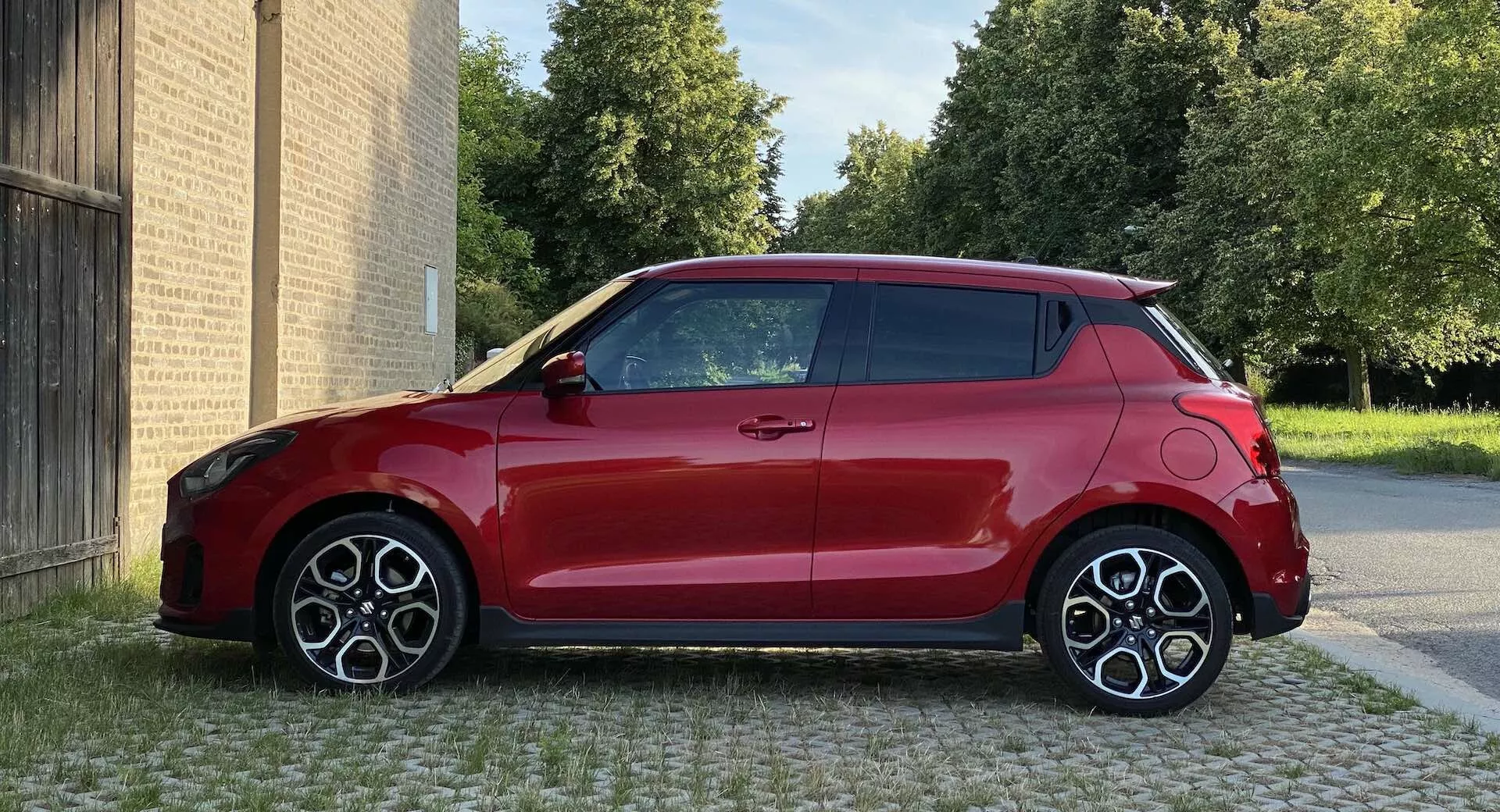 Imagen de un hatchback Suzuki Swift Sport 2017 rojo, tomada desde un ángulo de perfil lateral a nivel de los ojos, mostrando el lado derecho con un diseño bicolor de carrocería roja y techo negro. El coche está aparcado en una zona pavimentada con edificios al fondo y árboles visibles detrás de los edificios. El diseño compacto y moderno presenta ruedas grandes y deportivas de múltiples radios, líneas aerodinámicas visibles y una antena de aleta de tiburón. La iluminación es de luz del día, con calidad de imagen nítida y reflejos de superficie discernibles.