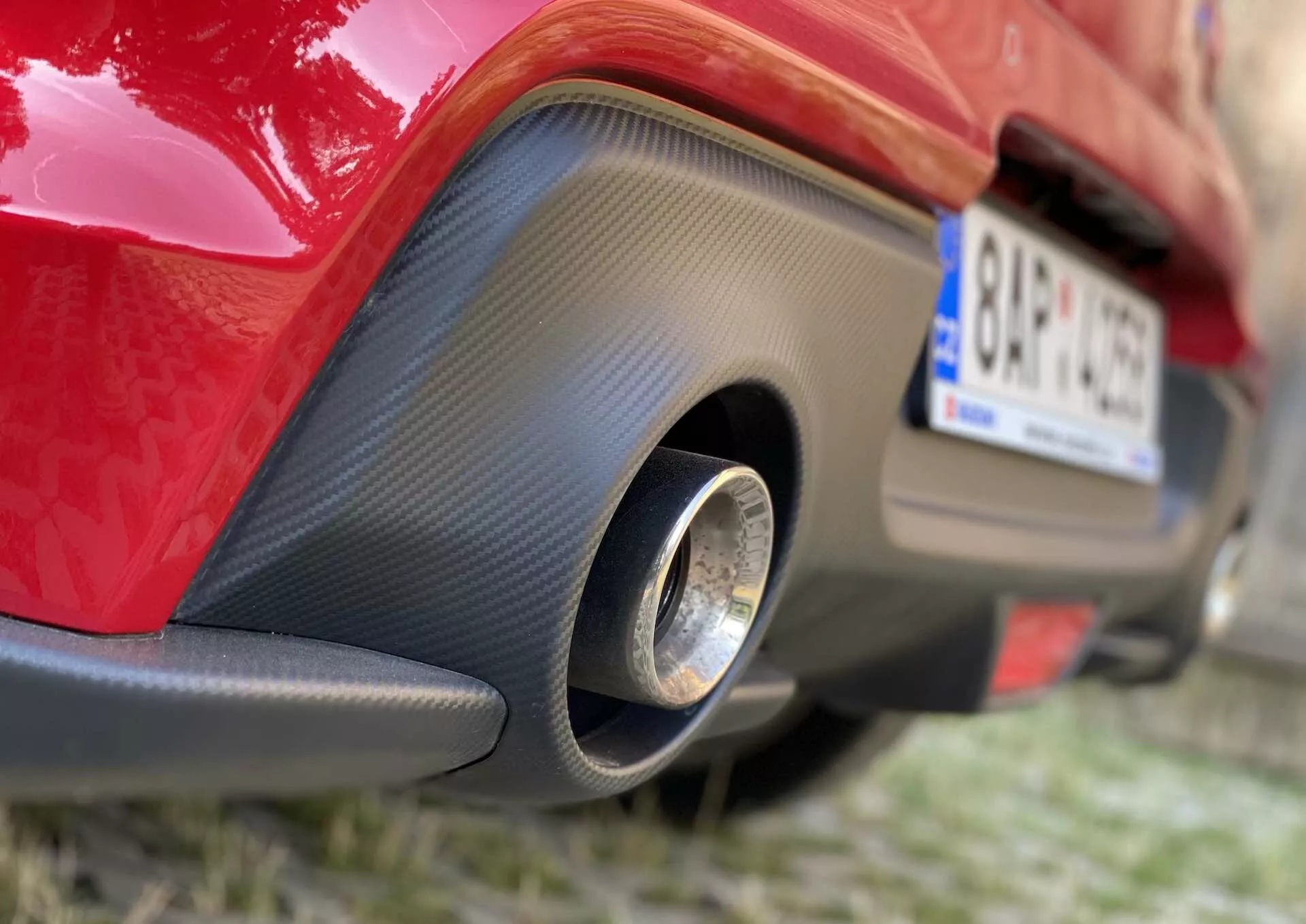 Primer plano de la sección trasera de un Suzuki Swift Sport rojo de 2017, destacando la parte inferior del parachoques trasero. La foto captura una vista en ángulo bajo desde la derecha, resaltando un inserto negro parecido a la fibra de carbono con un tubo de escape redondo de metal en el centro.