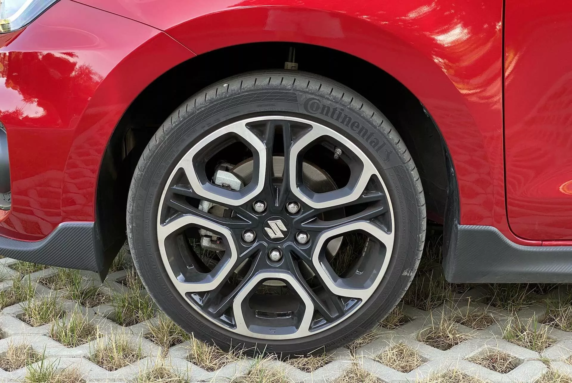 Primer plano de la rueda de un Suzuki Swift 2017 Swift Sport rojo, mostrando una llanta de aleación moderna con un diseño complejo de múltiples radios en acabados negro y plateado mecanizado. El neumático visible es un Continental PremiumContact, indicando alta calidad y buen estado del neumático. La carrocería del coche, brillante y bien mantenida, se extiende sobre la rueda, destacando las líneas estéticas del guardabarros y posiblemente un acabado deportivo. El vehículo está estacionado sobre adoquines hexagonales ecológicos que permiten el crecimiento de la hierba, enfatizando soluciones de estacionamiento sostenibles.