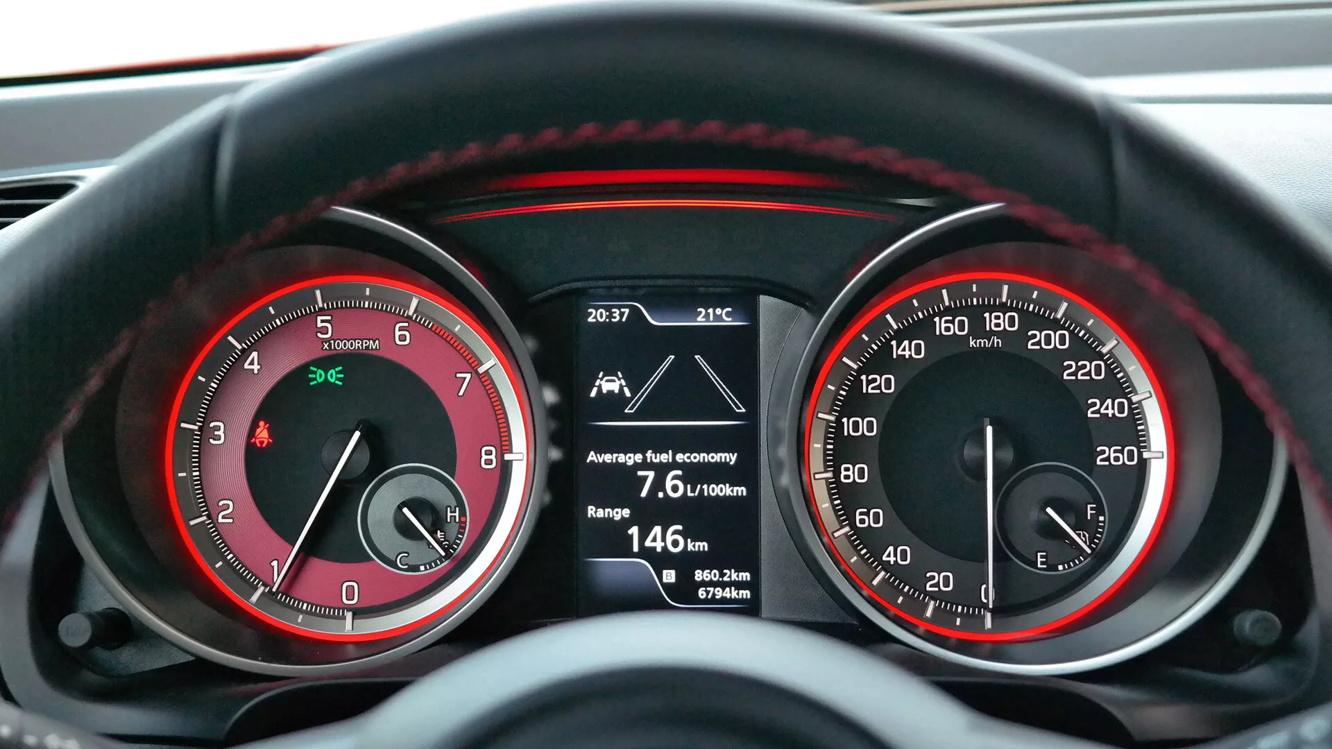 Una vista detallada y en primera persona del panel de instrumentos en un Suzuki Swift 2017 (Sport) mostrando un tacómetro analógico (hasta 7,000 RPM) y un velocímetro (hasta 260 km/h) con acentos rojos, y una pantalla digital central que indica una hora de 20:37, una temperatura exterior de 21°C, un promedio de consumo de combustible de 7.6L/100km, un rango restante de 146 km, dos odómetros de viaje (0.0 km y 367.6 km) y un kilometraje total de 80,622 km. La iluminación sugiere que el vehículo está operativo, probablemente en un ambiente con poca luz, reflejando una combinación de tecnología analógica y digital del tablero.