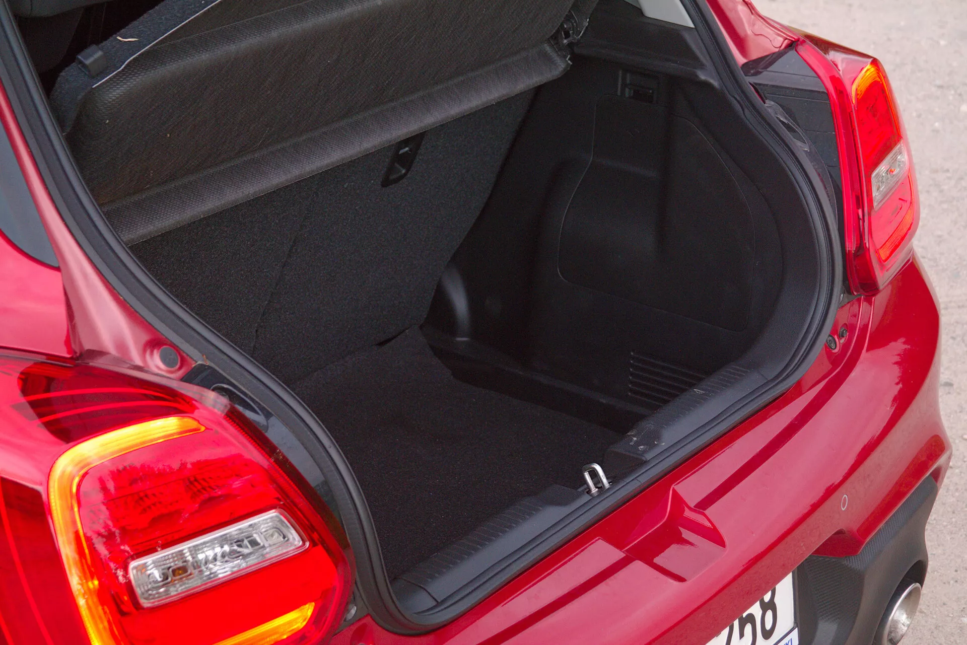 Vista en primer plano y en ángulo alto de la parte trasera de un Suzuki Swift Sport rojo de 2017, enfocándose en el área del maletero abierto. La toma está hecha desde el cuarto trasero derecho, mostrando el interior del maletero, la tapa del maletero y el diseño de las luces traseras del coche compacto. Las luces de freno están bajo una cubierta roja, mientras que las luces de reversa e indicadores de dirección están bajo una cubierta transparente. El maletero revela características estándar como pestillos y bisagras visibles, y alfombrado en el fondo y los lados. El borde bajo de carga sugiere una fácil manipulación del cargamento.