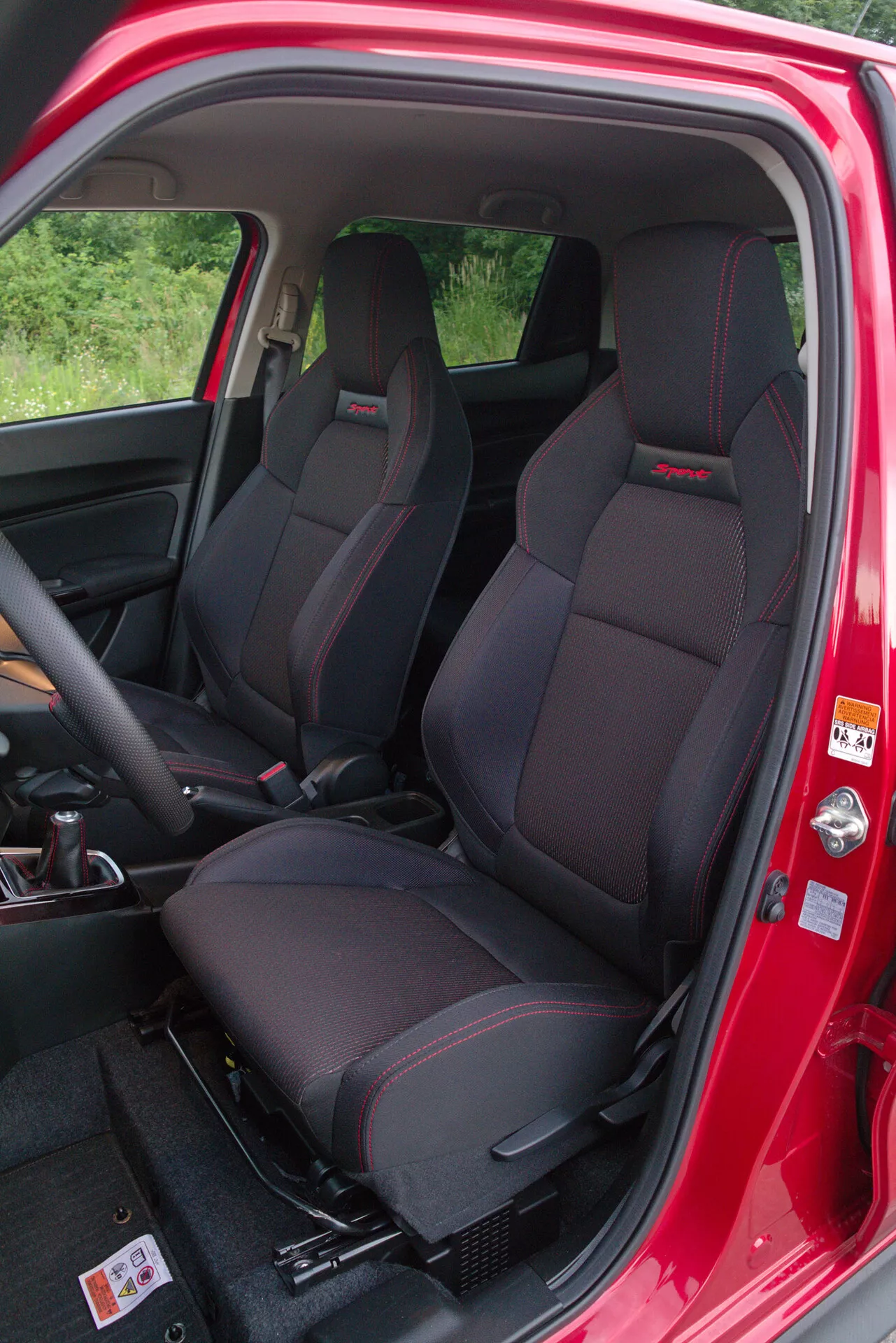 Vista interior del Suzuki Swift Sport 2017 desde el lado del pasajero, destacando los asientos delanteros tipo bucket con reposacabezas integrados y costuras rojas, puerta abierta del pasajero con pintura exterior roja y etiqueta de advertencia, panel de la puerta interior visible con controles y bolsillo en la puerta, visión parcial del tablero y volante, y alfombra oscura con umbral de plástico duradero.