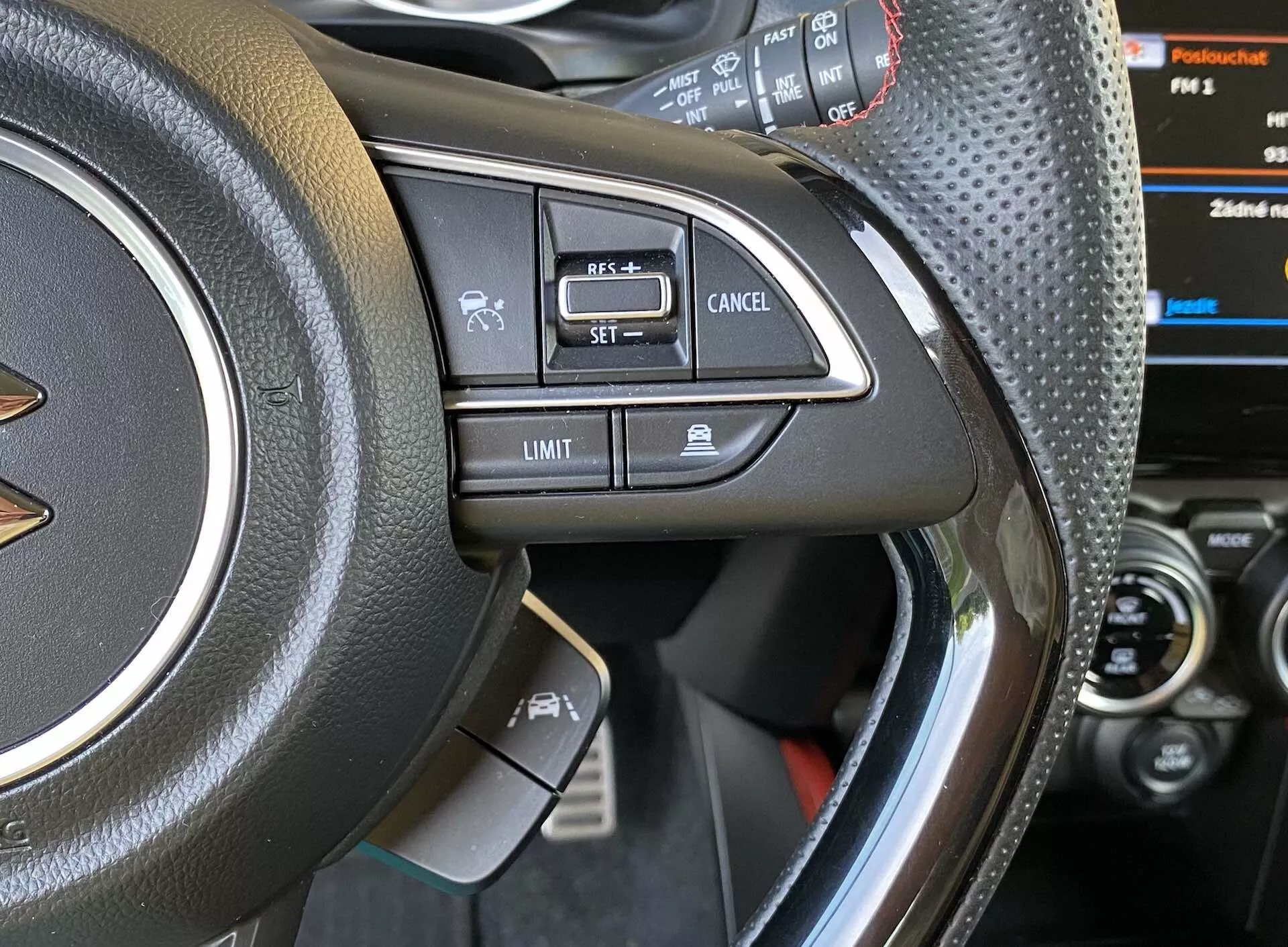 Vista de cerca del lado izquierdo del volante del Suzuki Swift 2017 Swift-Sport, mostrando los controles montados. Foto tomada desde el asiento del conductor o ligeramente fuera de la puerta del conductor, enfocándose en material de cuero o similar con costuras, acento metálico y varios botones como comando de voz, llamadas manos libres por Bluetooth, control de crucero 'SET,' 'Cancelar' y limitador de velocidad 'Límite.' Panel de instrumentos parcialmente visible con pantalla digital y consola central del automóvil.