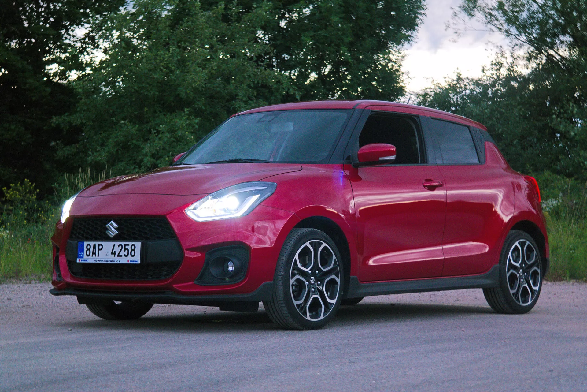 Vista frontal de tres cuartos de un coche hatchback rojo, que muestra elementos de diseño moderno como una parrilla frontal prominente, faros angulares elegantes probablemente con tecnología LED, llantas de aleación de múltiples radios y una línea de contorno dinámica a lo largo del perfil lateral. El vehículo tiene manijas de puertas del color de la carrocería y señales de giro integradas en los espejos laterales. La pintura roja brillante es uniforme y sin daños, estacionado en una superficie de asfalto con un fondo de vegetación, lo que indica un entorno rural o suburbano.