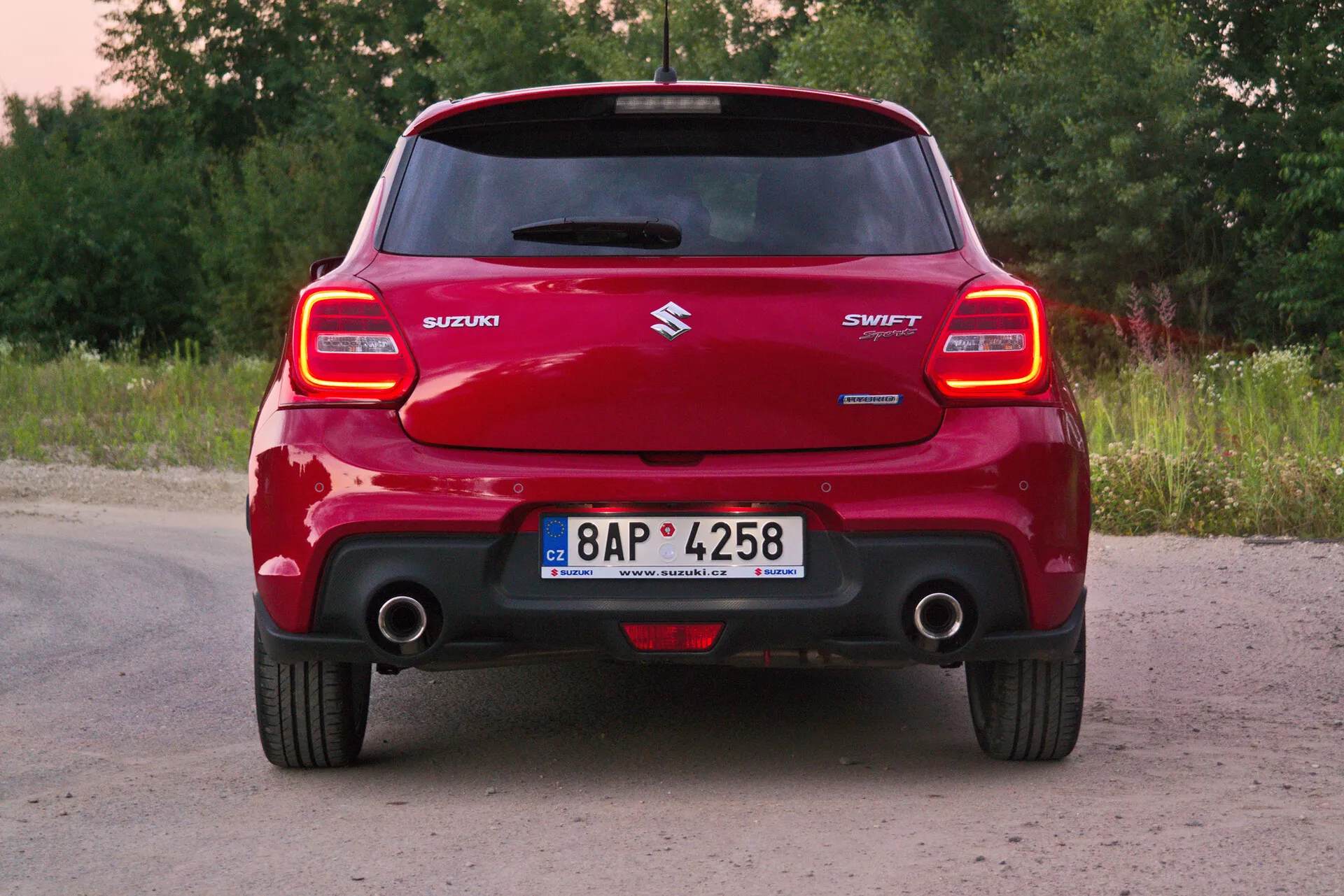 Vista trasera de un coche Suzuki Swift Sport 2017 rojo, enfocada en su ancho completo y parachoques trasero prominente, distintivos conjuntos de luces traseras, alerón de techo, logotipo central de Suzuki, nombre del modelo 'SWIFT', tubo de escape visible y matrícula en un entorno rural con vegetación y árboles.