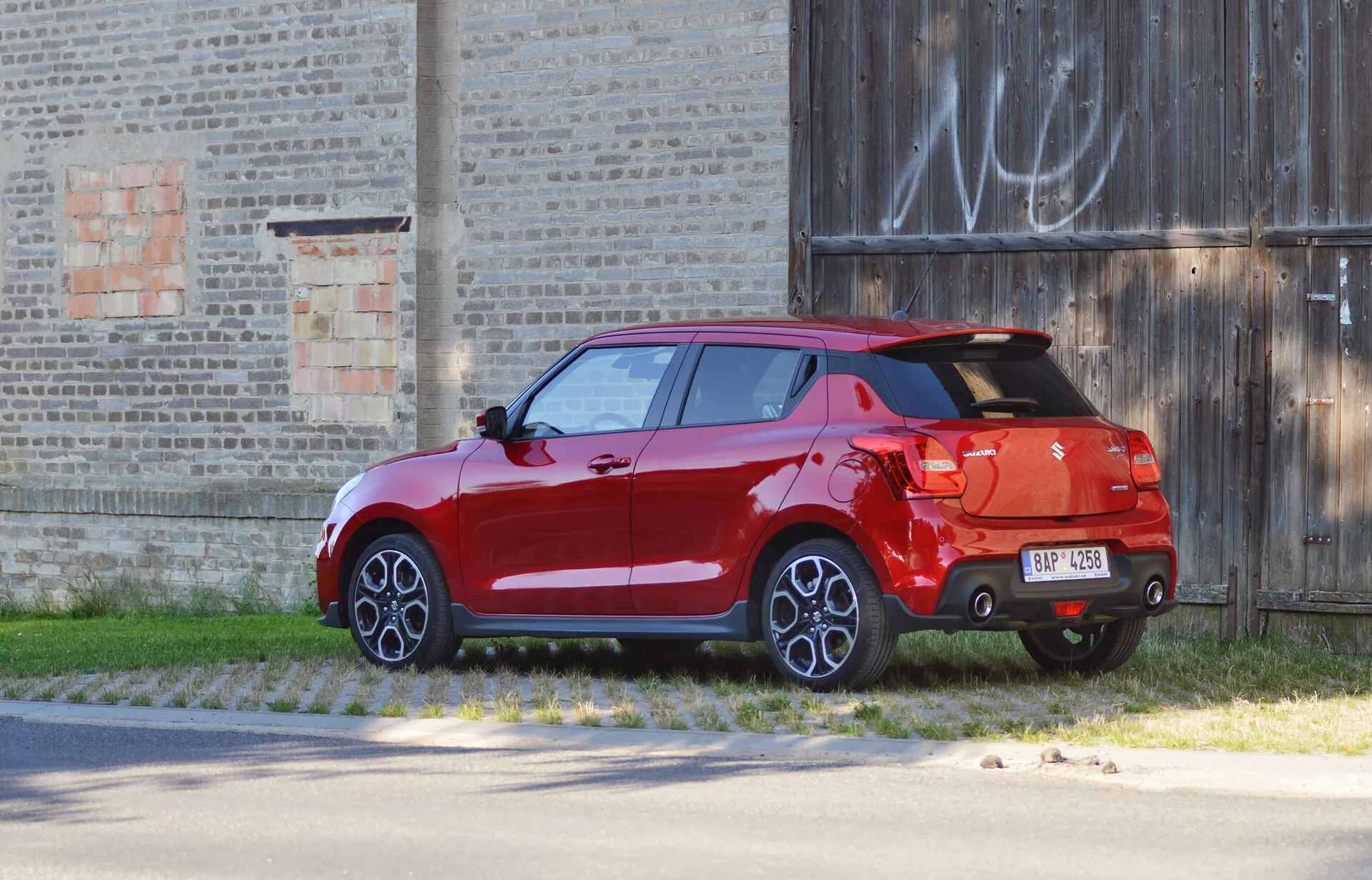Suzuki Swift Sport hatchback 2017 en color rojo brillante, capturado desde una vista trasera de tres cuartos, estacionado contra un fondo de un edificio viejo. La imagen resalta características de diseño como luces traseras, tapa trasera, destacado alerón trasero, parachoques bicolor con difusor, única salida de escape visible, llantas de aleación de múltiples radios, carrocería completamente pintada, incluidos los espejos laterales y manillas de las puertas, indicando un nivel de equipamiento superior o opciones de lujo.
