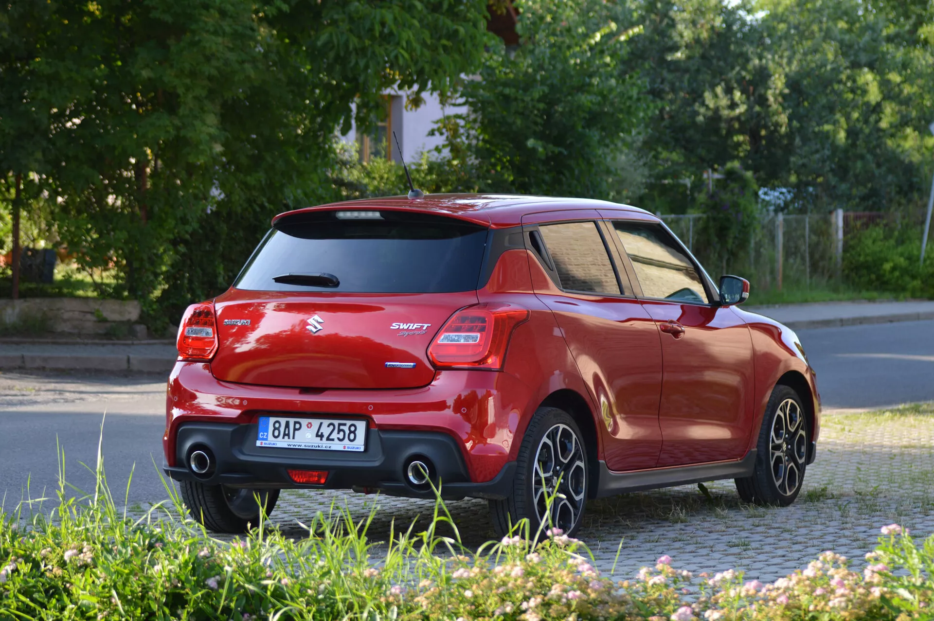 La imagen muestra un coche hatchback Suzuki Swift Sport 2017 de color rojo vibrante capturado desde un ángulo de tres cuartos trasero, mostrando el lateral y la parte trasera. Destacan el techo y los pilares negros en contraste, un alerón trasero elevado, luces traseras distintivas y una insignia de modelo 'Swift'. La fotografía tomada desde nivel del suelo muestra las llantas de aleación de cinco radios en movimiento, sugiriendo que el coche está en marcha. La iluminación indica que es temprano por la mañana o al final de la tarde, con sombras largas.