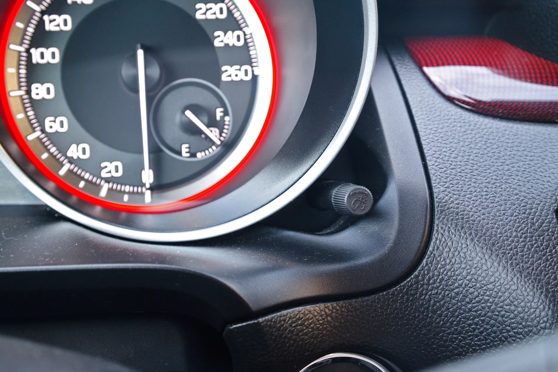 Vista de cerca del cuadro de instrumentos de un Suzuki Swift Sport 2017, destacando el velocímetro y el indicador de combustible. El velocímetro tiene números blancos en incrementos de 20, alcanzando hasta 260 km/h o mph, con la aguja en cero. El diseño presenta un tema deportivo en rojo y blanco con un círculo rojo en la circunferencia interior del dial. El indicador de combustible, casi lleno, está a la derecha. La toma está en ángulo desde el asiento del conductor con una textura granulada negra alrededor de los indicadores y un interior de puerta rojo y negro a juego, posiblemente de cuero y plástico. Hay un pequeño objeto circular cerca del velocímetro.