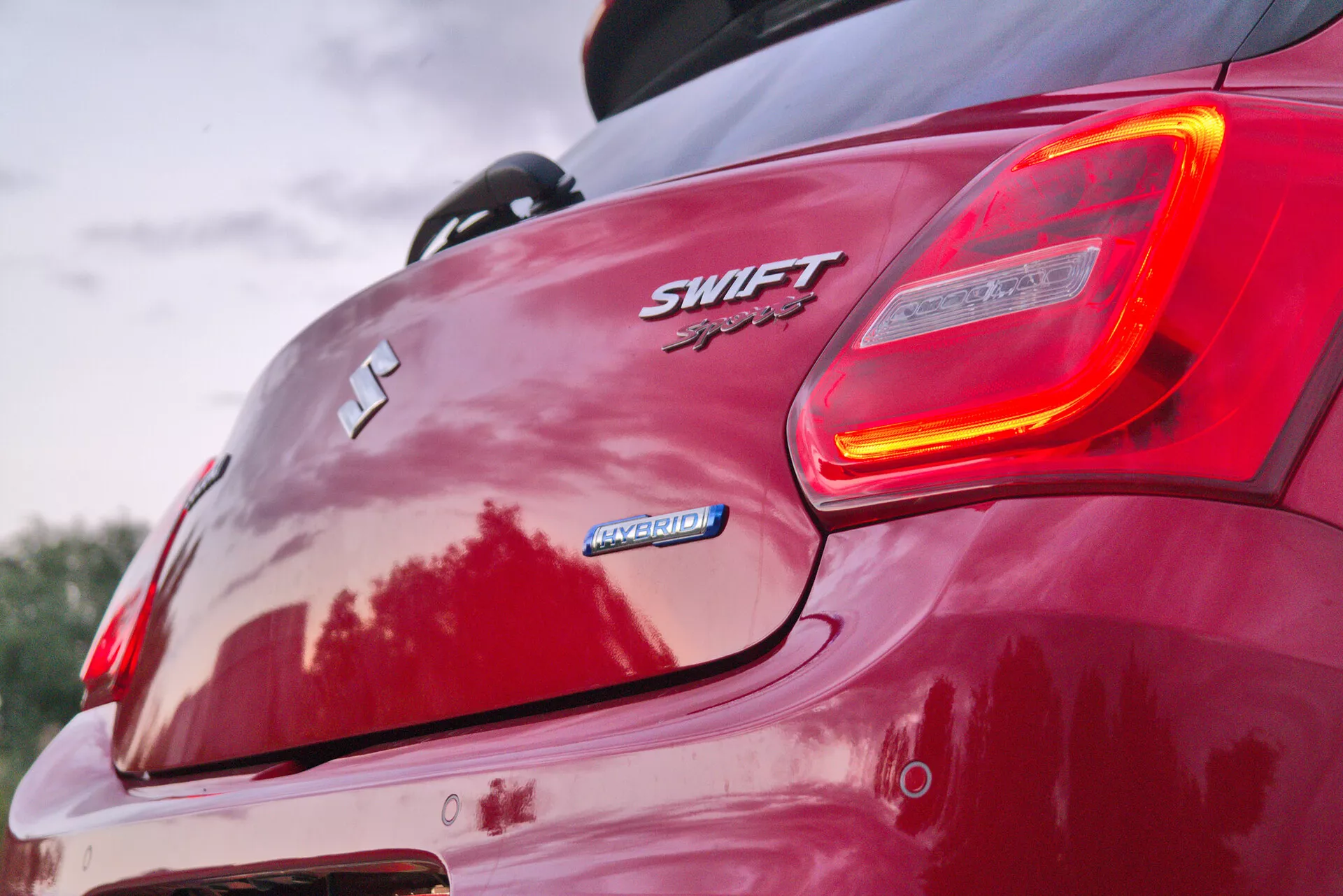 Esta imagen en primer plano captura la esquina trasera derecha de un Suzuki Swift Sport 2017 rojo, enfocándose en el emblema y la luz trasera iluminada del coche. Desde un ángulo bajo, acentúa los contornos traseros del coche, el spoiler y la estética deportiva. Los elementos visibles incluyen el emblema 'S' de Suzuki, las insignias 'SWIFT' y 'Sport', la insignia 'HYBRID', el diseño del parachoques trasero, las llantas de aleación y reflejos que sugieren un acabado de pintura brillante.