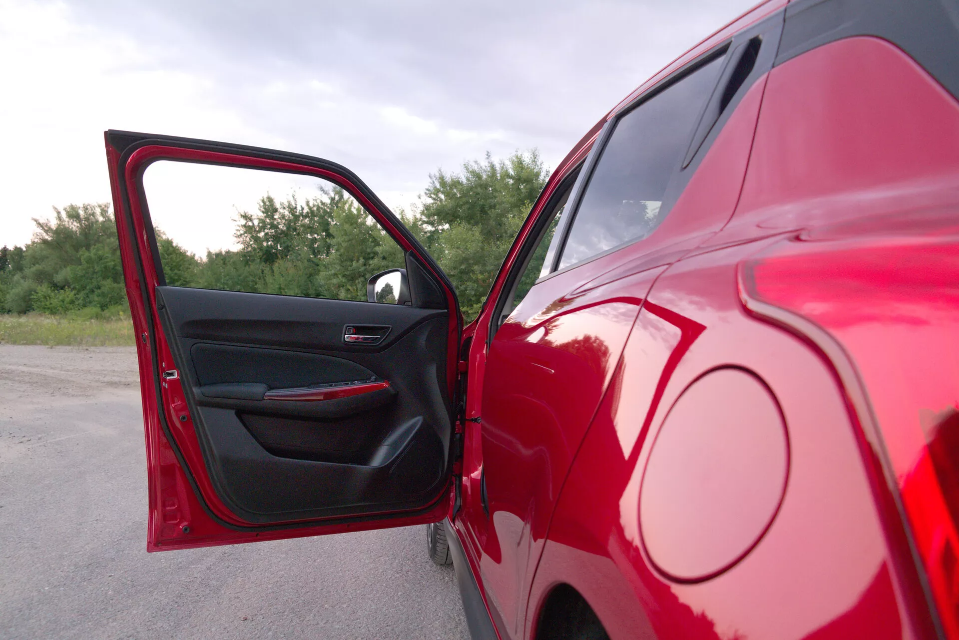 Vista lateral parcial de un Suzuki Swift Sport 2017 en rojo, capturada desde un ángulo de cerca a medio rango con la puerta del conductor abierta. La foto destaca la mitad trasera del vehículo estacionado, mostrando un exterior rojo brillante y un panel interior de puerta oscuro con controles de ventanas eléctricas y una manija de puerta elegante. El fondo presenta follaje natural, sugiriendo que el coche está estacionado al aire libre.