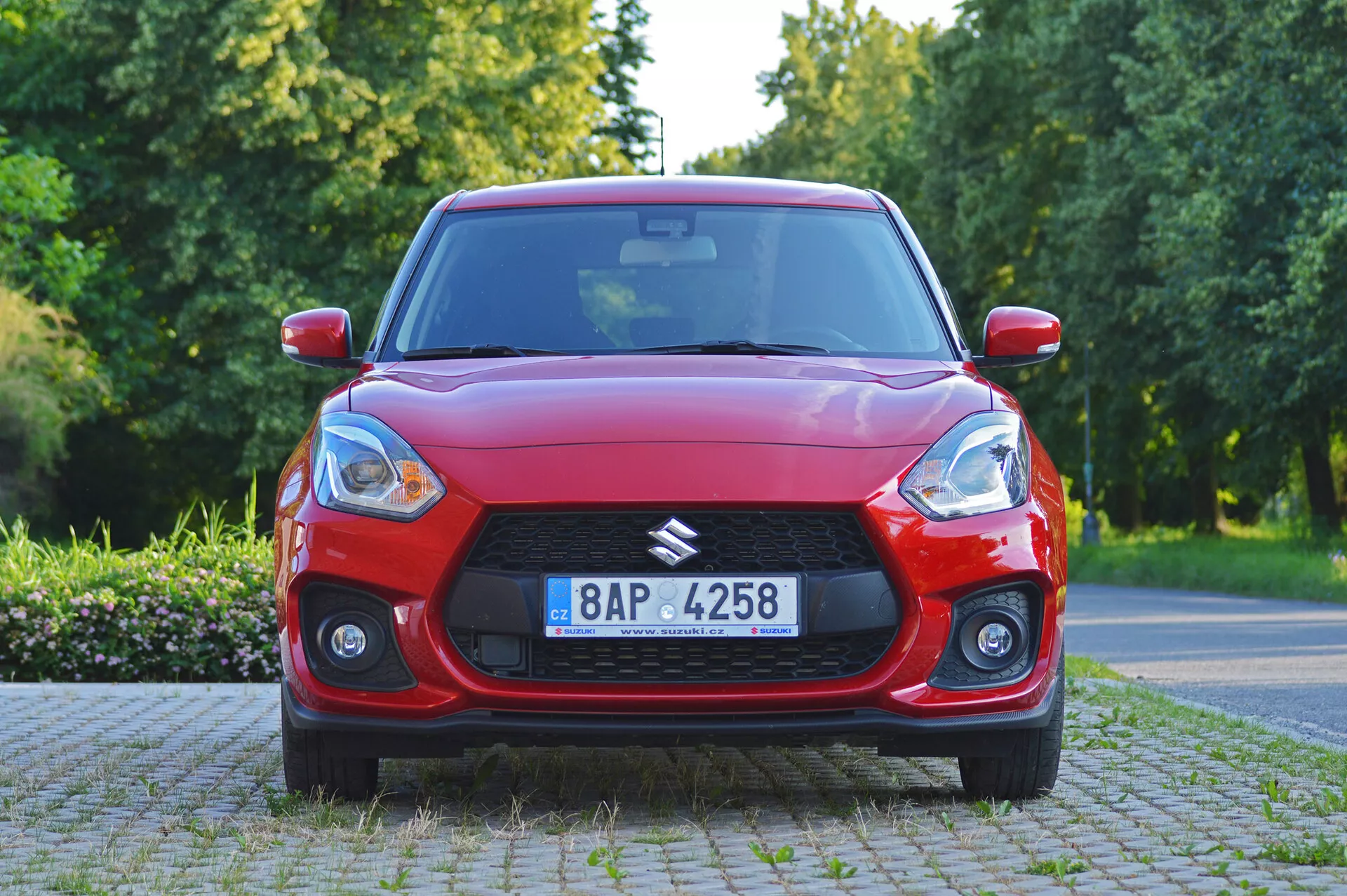 Vista de ángulo frontal de un hatchback rojo Suzuki Swift Sport 2017 con rejilla prominente y faros inclinados, con iluminación natural diurna. El capó del coche muestra líneas aerodinámicas, con faros antiniebla que mejoran su anchura y deportividad. Detalles incluyen un posible dispositivo tecnológico en el parabrisas, llantas de aleación multifradio y una placa de matrícula visible. El vehículo está estacionado en un entorno suburbano o parque con árboles y vegetación.