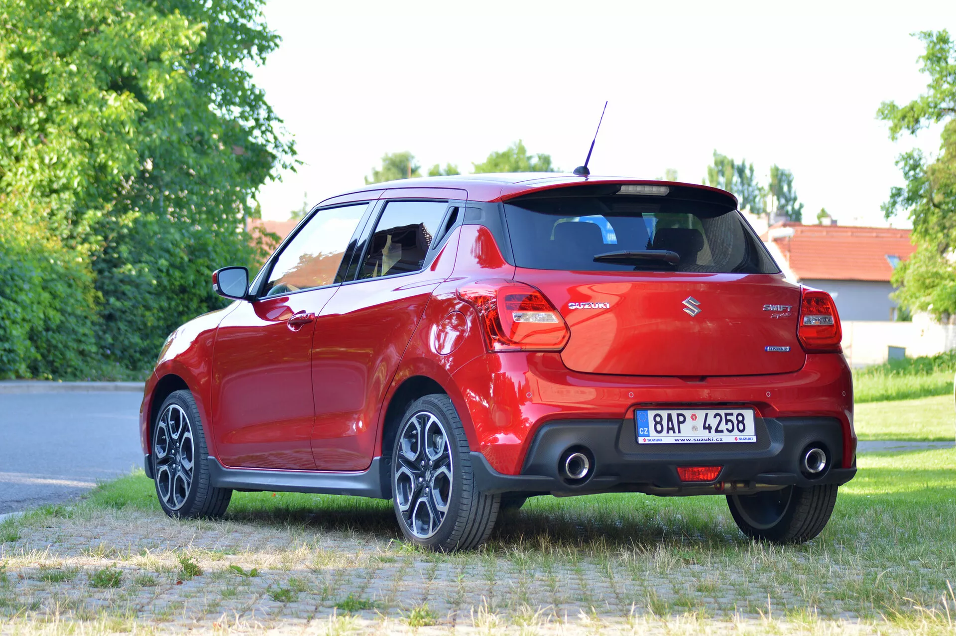 Imagen detallada de un moderno hatchback rojo Suzuki Swift Sport 2017 colocado en una superficie pavimentada con césped y árboles en el fondo. La foto está tomada desde una perspectiva de tres cuartos traseros, mostrando el lado trasero derecho del coche. Las características visibles incluyen las luces traseras, el parabrisas trasero, el parachoques, el sistema de escape doble, las llantas de aleación multirrayos y una rueda trasera girada. La matrícula está difuminada para mantener la privacidad.