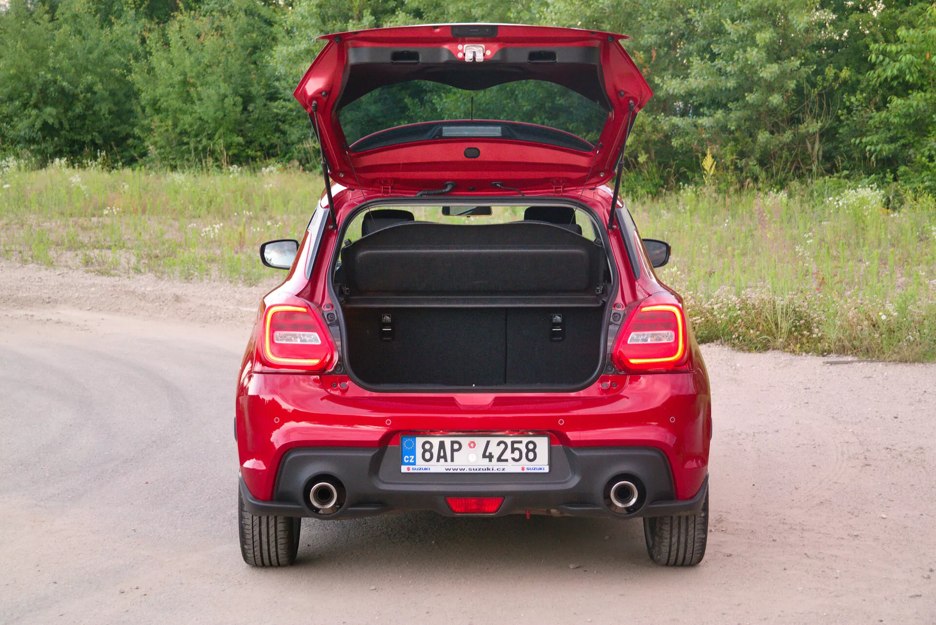 Vista trasera de un Suzuki Swift Sport 2017 rojo con la tapa del maletero abierta, mostrando características traseras simétricas, luces traseras, escape visible en el lado del pasajero y limpiaparabrisas trasero. El vehículo está estacionado en un camino sin pavimentar con pasto y vegetación en el fondo. La matrícula está montada en el centro del parachoques trasero. El maletero vacío revela la capacidad de almacenamiento y el revestimiento interior, con una manija en la parte interna de la tapa para un uso ergonómico. Los asientos traseros parcialmente plegados sugieren una división 60/40 para un espacio de carga ampliable. El parachoques trasero simple indica características estándar sin complementos tecnológicos avanzados.