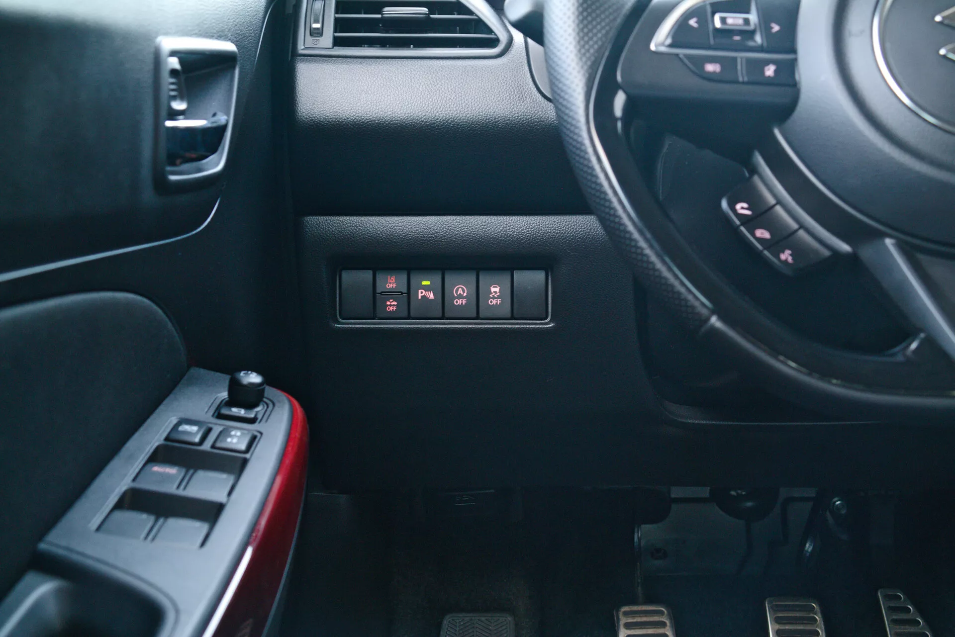 Vista interior de un Suzuki Swift Sport 2017 desde la perspectiva del pasajero, mostrando el tablero del conductor y el lado de la consola central. Incluye vista parcial del volante con botones multifuncionales, tablero con botones de pulsar, puerta del conductor con controles de ventanas eléctricas y espejos, pedales metálicos deportivos para una transmisión manual y texturas y materiales detallados.