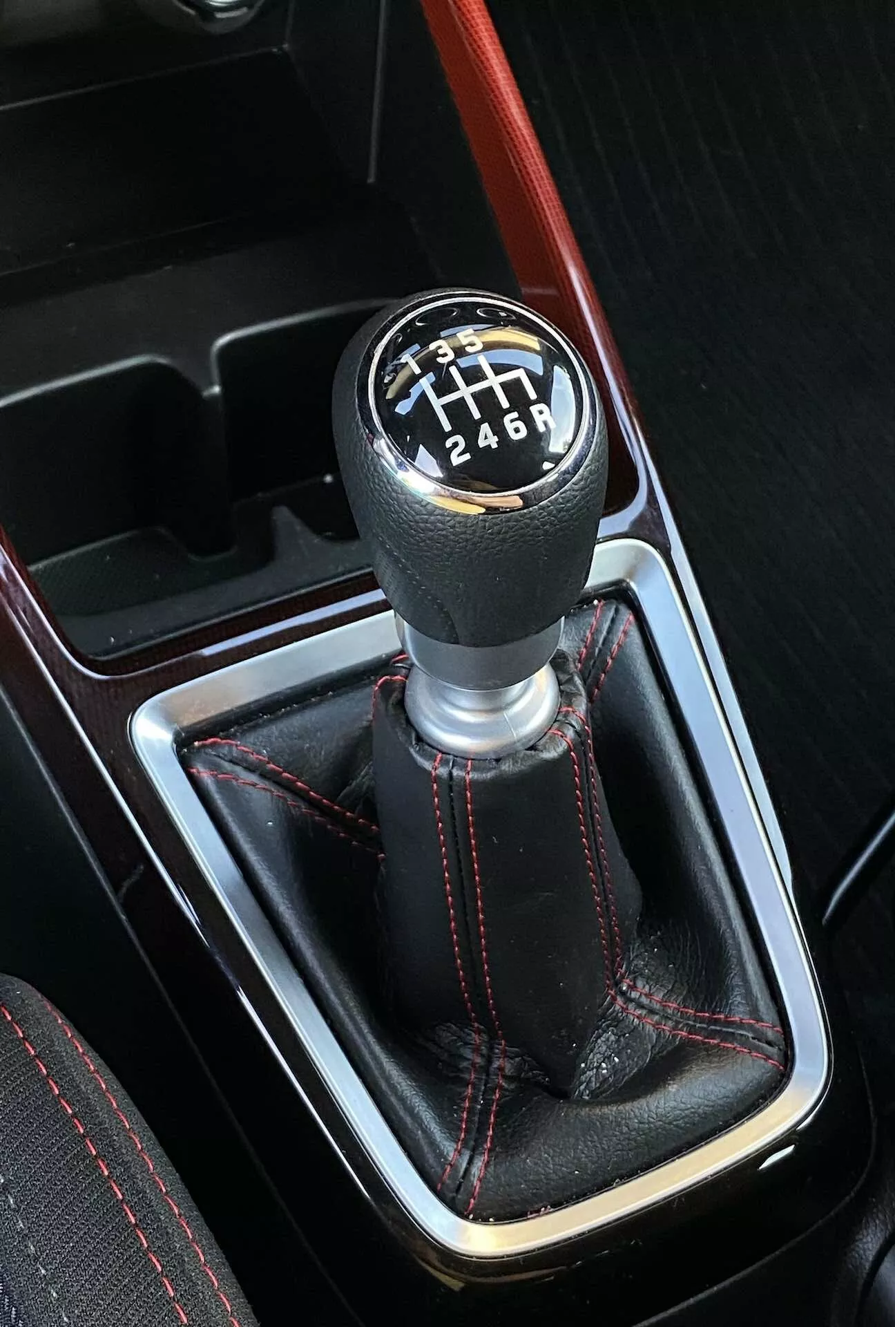 Vista superior, ligeramente angulada de la palanca de cambios de transmisión manual en un Suzuki Swift Sport 2017, con un patrón de seis velocidades delineado en blanco sobre un fondo negro. La palanca de cambios está situada en una consola central, rodeada por una funda de cuero con costuras de contraste rojas y una línea de acento roja que corre paralela a la palanca de cambios. La imagen muestra un interior deportivo con destellos de un embellecedor de imitación de madera, indicando elementos de diseño de lujo.