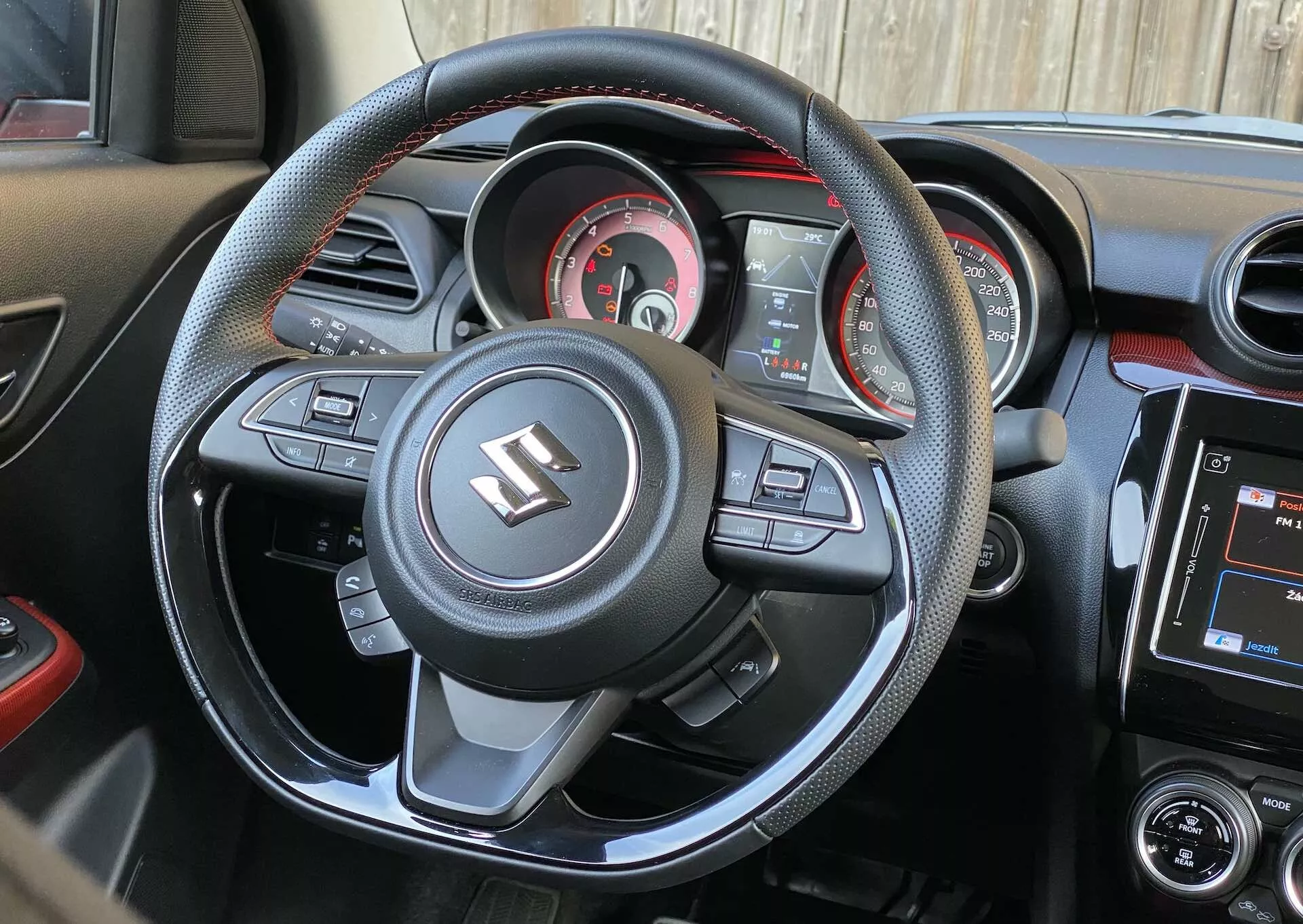 Vista del interior de un Suzuki Swift Sport 2017 desde la perspectiva del asiento del conductor, mostrando el volante deportivo de tres radios con parte inferior plana y botones multifuncionales para tecnología multimedia y de conveniencia, incluidos ajuste de volumen, comandos de voz, navegación por el menú y control de crucero. El volante está envuelto en cuero con detalles de costura en contraste y presenta prominentemente el emblema de Suzuki. Detrás, el conjunto de instrumentos muestra un velocímetro analógico central, un tacómetro, un indicador de combustible y una pequeña pantalla digital para datos adicionales del vehículo. El tablero de instrumentos integra ventilas de aire y una pantalla táctil de infoentretenimiento con controles de audio y aire acondicionado. El esquema de colores es predominantemente negro con adornos metálicos y acentos rojos, subrayando una estética deportiva.