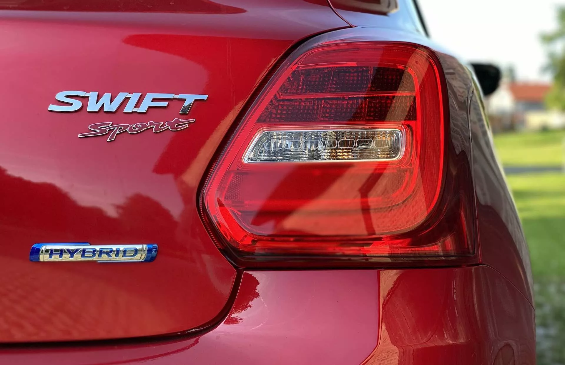 Vista de cerca de la parte trasera de un Suzuki Swift Sport rojo 2017, enfocada en el conjunto de luces traseras LED del lado del conductor. La imagen captura el acabado brillante de la pintura del coche, las insignias SWIFT Sport y HYBRID, y los contornos del guardabarros trasero y parte de la tapa del maletero. El enfoque superficial difumina el fondo y resalta los detalles del conjunto de luces e insignias, destacando el diseño moderno y la tecnología híbrida del coche.