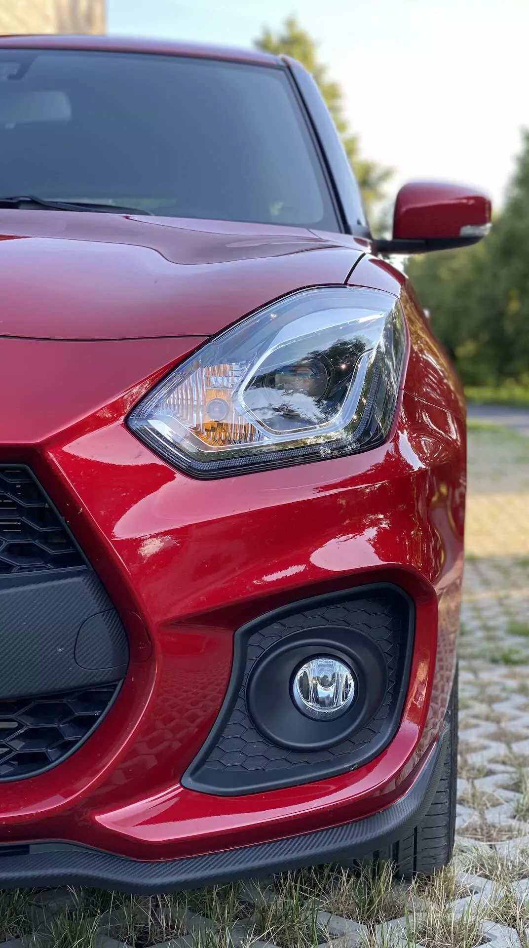 Vista de cerca de un Suzuki Swift Sport rojo 2017 desde un ángulo bajo, centrada en la parte delantera izquierda con el faro del lado del conductor de diseño intrincado que posiblemente incluye tecnología de iluminación LED o halógena, emblema parcialmente desenfocado en la parrilla y espejo lateral aerodinámico en un fondo natural con vegetación.
