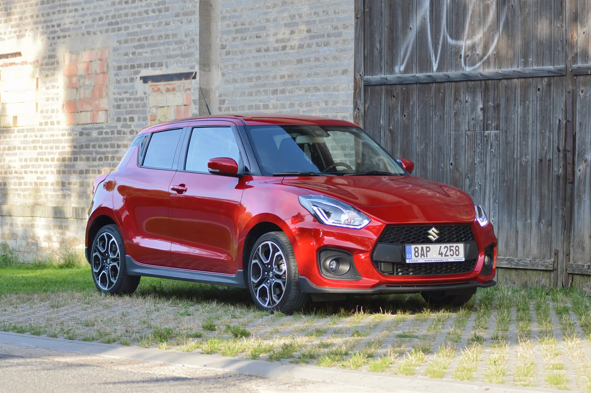 Imagen de un hatchback Suzuki Swift Sport 2017 rojo capturado desde un ángulo frontal de tres cuartos, destacando los perfiles frontal y lateral. Las características de diseño del coche incluyen una parrilla frontal prominente con un logo integrado, faros elegantes, carcasas de luces antiniebla y llantas de aleación de múltiples radios. La toma está hecha desde una altura ligera, proporcionando una vista de arriba hacia abajo del capó y el techo. El entorno incluye un primer plano de hierba y una pared de ladrillo con graffiti en el fondo, enfatizando la apariencia deportiva y dinámica del vehículo.