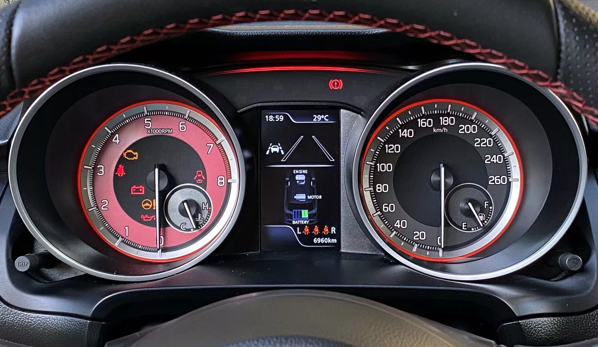 Vista de cerca del panel de instrumentos en un Suzuki Swift Sport 2017 desde la perspectiva del asiento del conductor. La imagen muestra el diseño de doble medidor con un cuentarrevoluciones rojo (hasta 8,000 RPM) a la izquierda y un velocímetro a juego con tema rojo (hasta 260 km/h) a la derecha, con acentos cromados y numeración blanca para una mejor visibilidad. Una pantalla digital pequeña central muestra la hora (18:59), la temperatura exterior (29°C), la selección de marcha atrás y la lectura del odómetro (96604 km). Las luces de advertencia adicionales están ubicadas en el borde superior, junto con costuras rojas que añaden un toque deportivo.