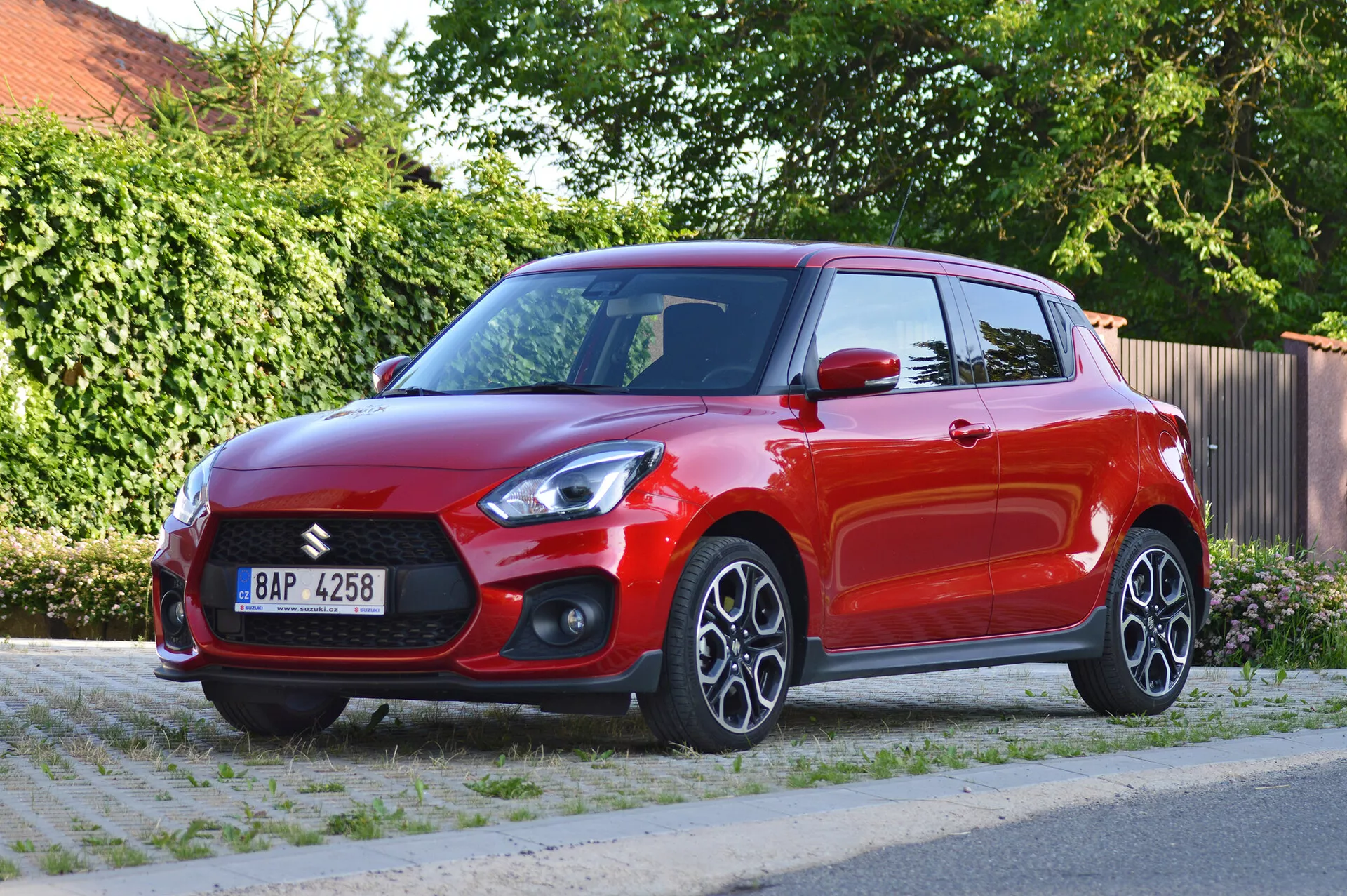La imagen muestra un vibrante hatchback Suzuki Swift Sport 2017 rojo, fotografiado desde una vista frontal de tres cuartos. Destaca la parrilla frontal prominente con el emblema de Suzuki, los faros LED angulares y nítidos, las carcasas de los faros antiniebla y las entradas de aire en el parachoques delantero inferior. El capó presenta líneas de contorno. El perfil lateral muestra llantas de aleación multirradio, señales de giro integradas en los espejos laterales del color de la carrocería y manijas de puertas del mismo color. La línea de hombros alta y la línea del techo ligeramente inclinada añaden una apariencia deportiva. La pintura brillante y las ventanas ligeramente tintadas indican un vehículo bien mantenido. La foto se toma durante el día con una vista clara de una calle residencial y un fondo de follaje verde, proporcionando una exposición equilibrada y destacando los elementos de diseño del coche.