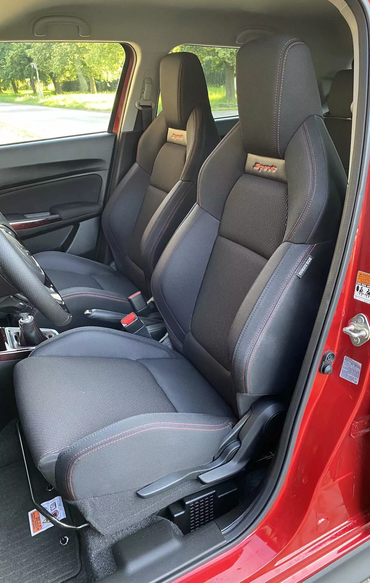 El interior de un Suzuki Swift Sport 2017 se muestra desde una perspectiva del lado del pasajero a través de la puerta delantera abierta. El enfoque está en los asientos delanteros modernamente contorneados y deportivos con reposacabezas integrados y soportes laterales prominentes, posiblemente con una combinación de cuero sintético y tela y costuras de contraste. En la parte superior de los respaldos se observan detalles de logotipos en relieve o bordados. Ambos asientos están equipados con un sistema de cinturón de seguridad de tres puntos, y el lado del conductor tiene un mecanismo ajustable para la altura del cinturón en el pilar B. La consola central entre los asientos tiene compartimentos de almacenamiento y una palanca de freno de mano. El panel de la puerta interior de color oscuro incluye una manija de la puerta, control de ajuste de espejos y controles de ventanas eléctricas. El piso está cubierto con una alfombra oscura y la base de los asientos muestra rieles de montaje. Las condiciones de luz diurna y el entorno natural exterior son visibles a través de las ventanas, proporcionando reflejos y sombras que mejoran los detalles de la cabina.