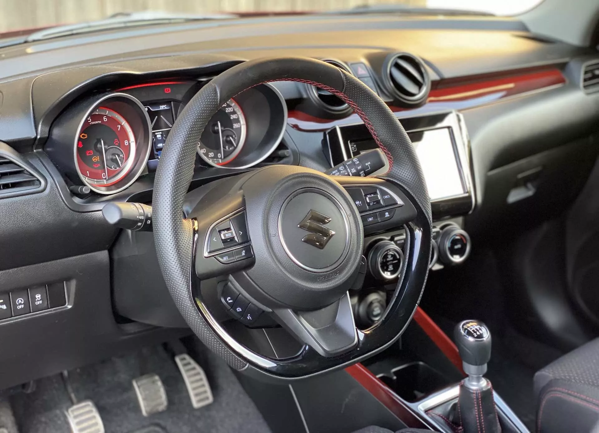 Vista interior de un Suzuki Swift Sport 2017 desde un ángulo ligeramente derecho, que presenta el volante multifuncional forrado en cuero, panel de instrumentos analógico con tacómetro y velocímetro, consola central con pantalla táctil multimedia y controles manuales, y palanca de cambios para transmisión manual.