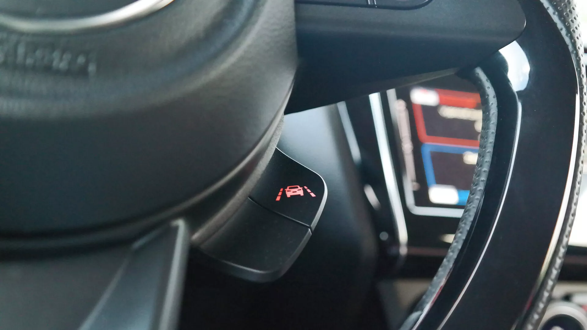 Vista del interior de un Suzuki Swift 2017 (Swift-sport) desde la perspectiva del asiento del conductor, centrada en el volante y la palanca de control. El volante de color oscuro, posiblemente de cuero, presenta un ribete metálico o brillante. Una palanca de control de crucero con un símbolo de un medidor y una flecha sobresale de la columna de dirección. En el fondo borroso, el panel de instrumentos digital muestra varios números y líneas, lo que indica características tecnológicas avanzadas. Los materiales del interior muestran una mezcla de texturas sintéticas o de cuero con costuras intrincadas, y la poca profundidad de campo resalta la palanca de control con el tablero de instrumentos desenfocado.